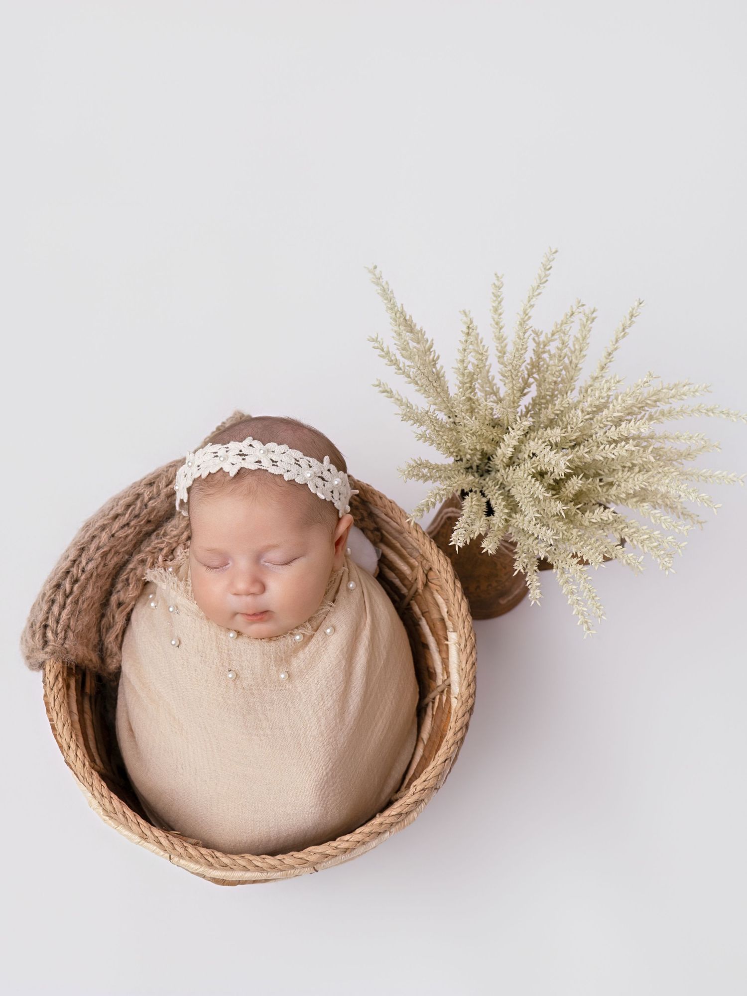 Sleeping newborn baby wrapped in cream swaddle inside woven basket with white plant.