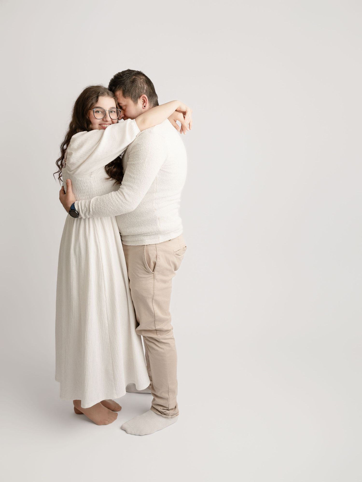 Couple sharing a warm embrace in matching white and beige outfits against a light background.