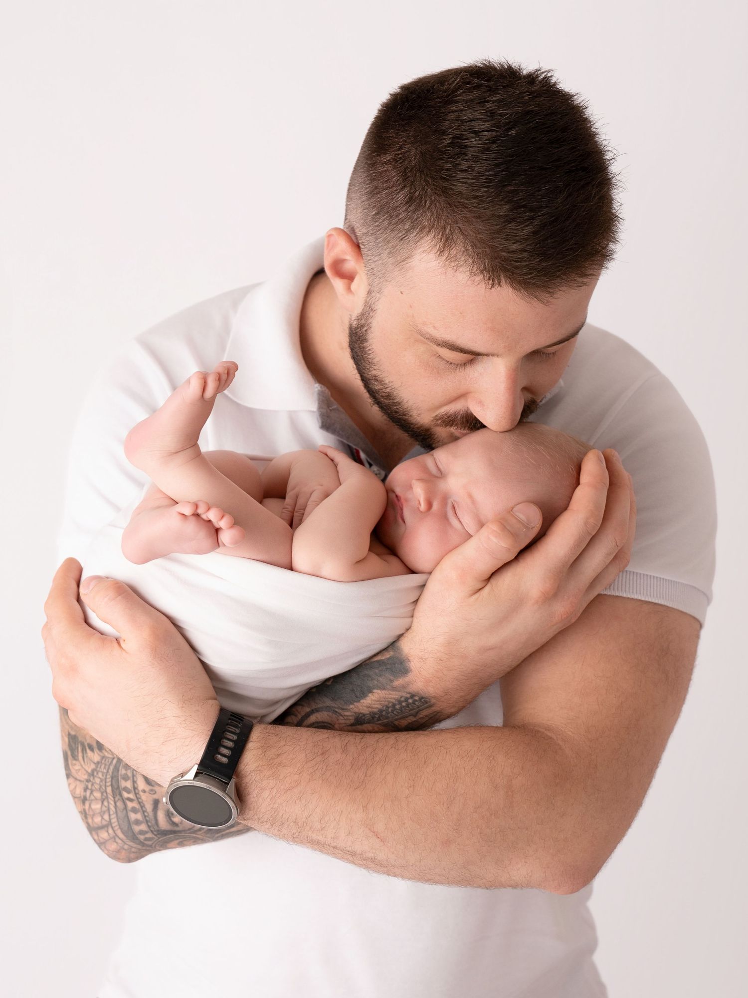 A father tenderly kisses his sleeping newborn baby, cradling the infant in his tattooed arms against a white background.