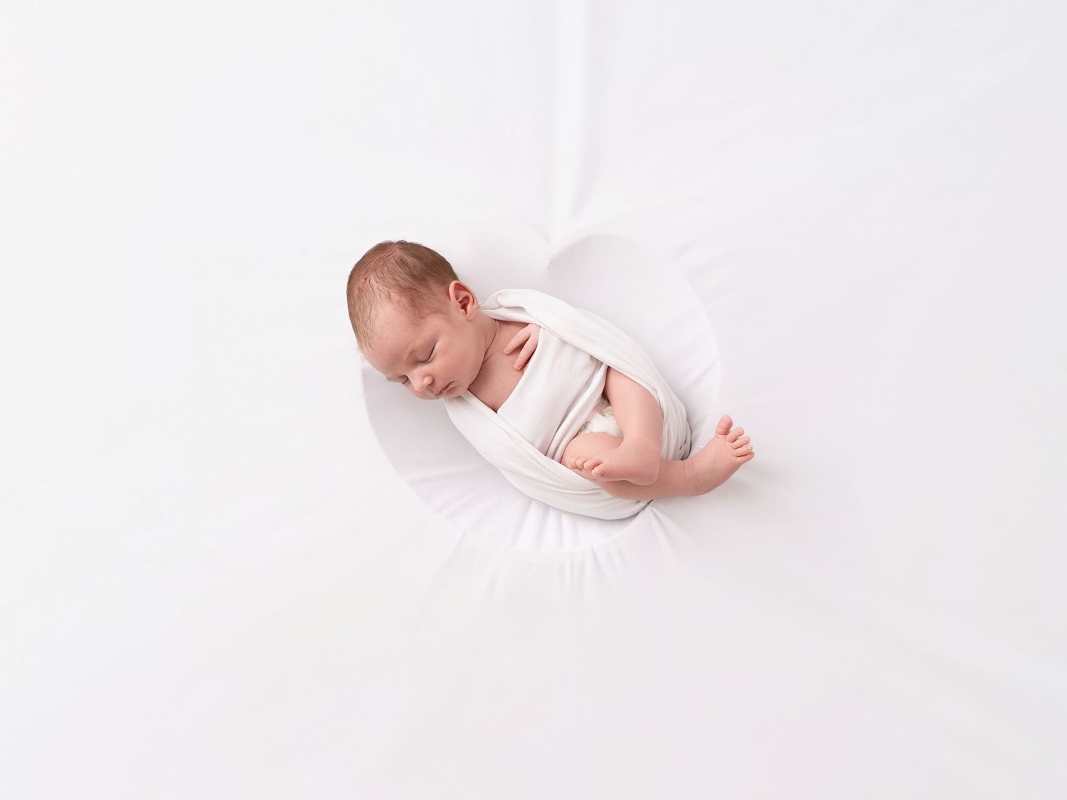 Newborn baby swaddled in white fabric sleeping peacefully on a soft white background in a professional photo.