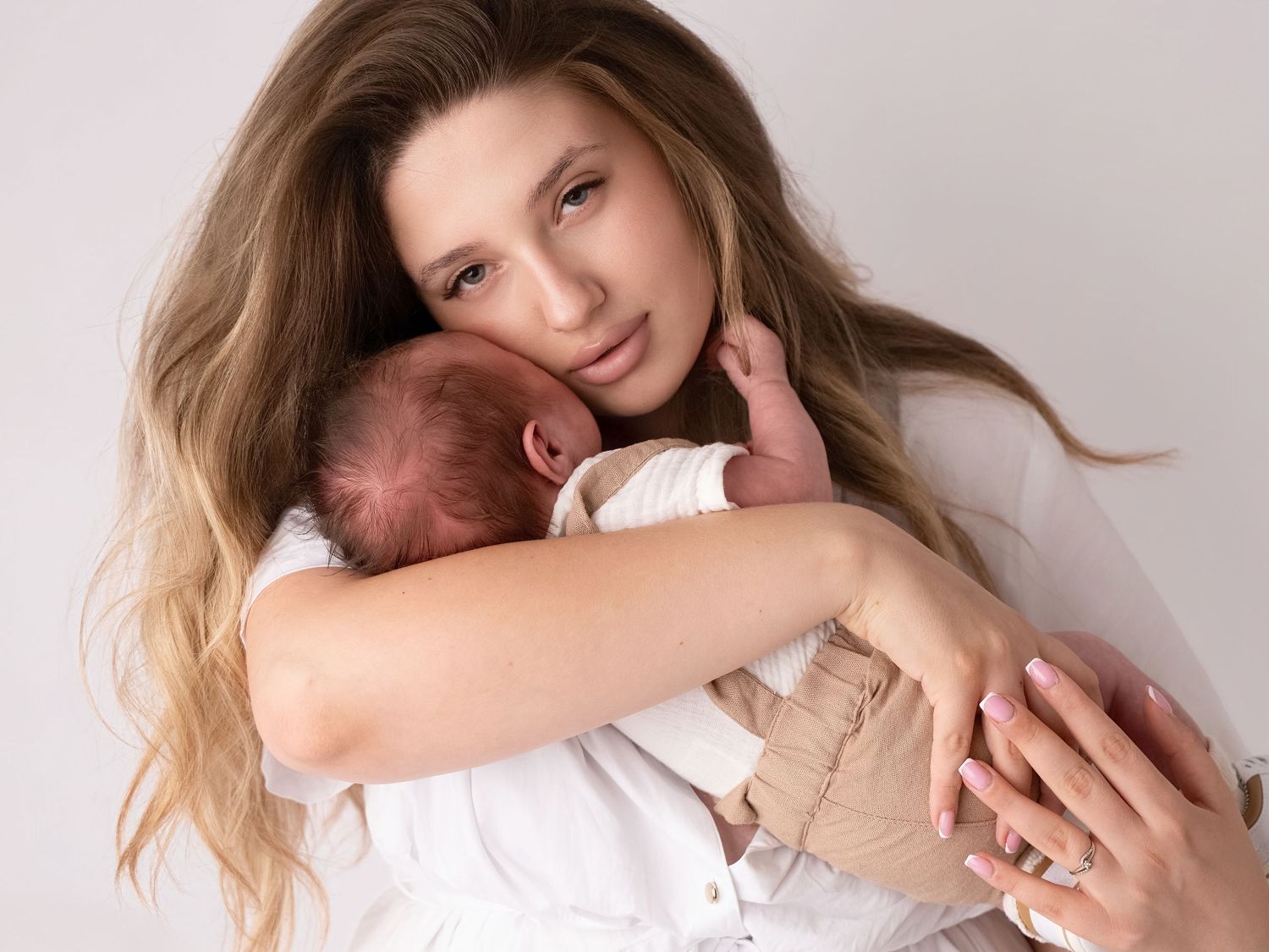 Young mother with long hair tenderly holds sleeping newborn baby against her chest in soft white studio setting.