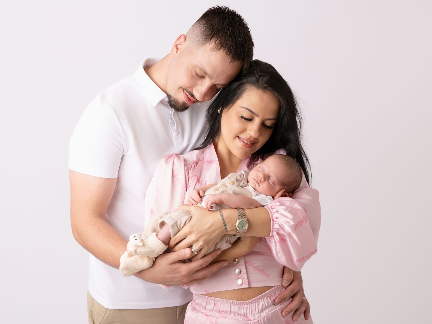 A couple in pink and white outfits tenderly holds their newborn baby against a soft white background.