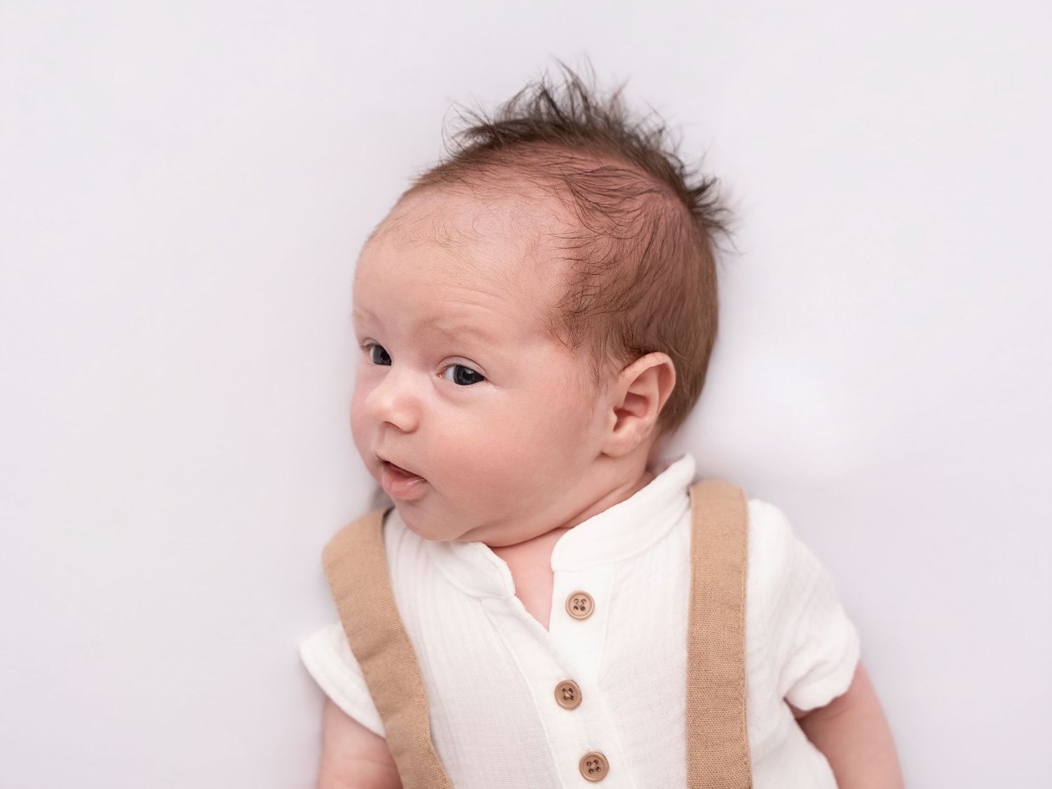 Adorable newborn baby in cream linen outfit with suspenders lying on white background looking sideways.
