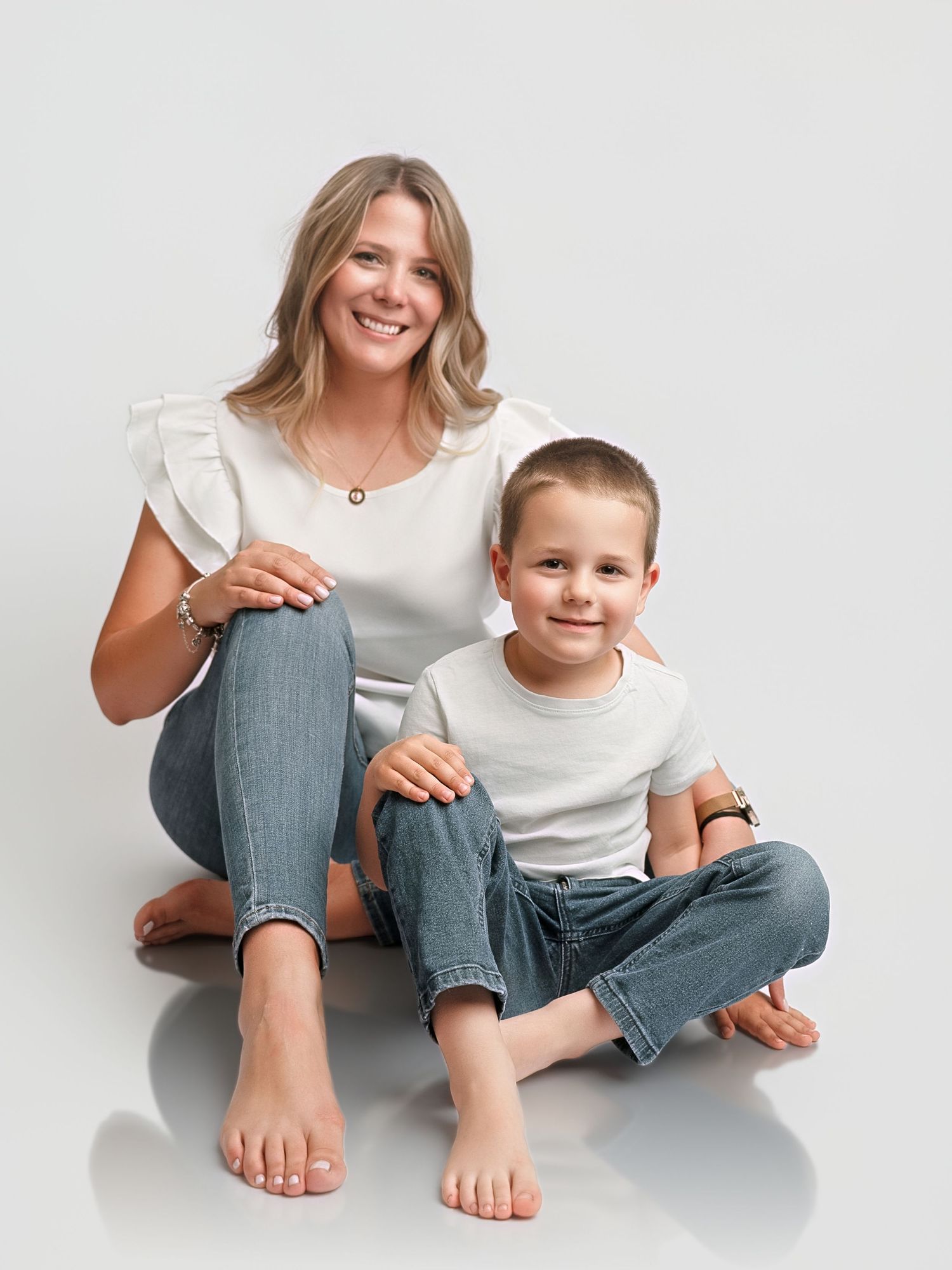 Smiling mother and young son in matching white tops and jeans sit together on white studio floor.