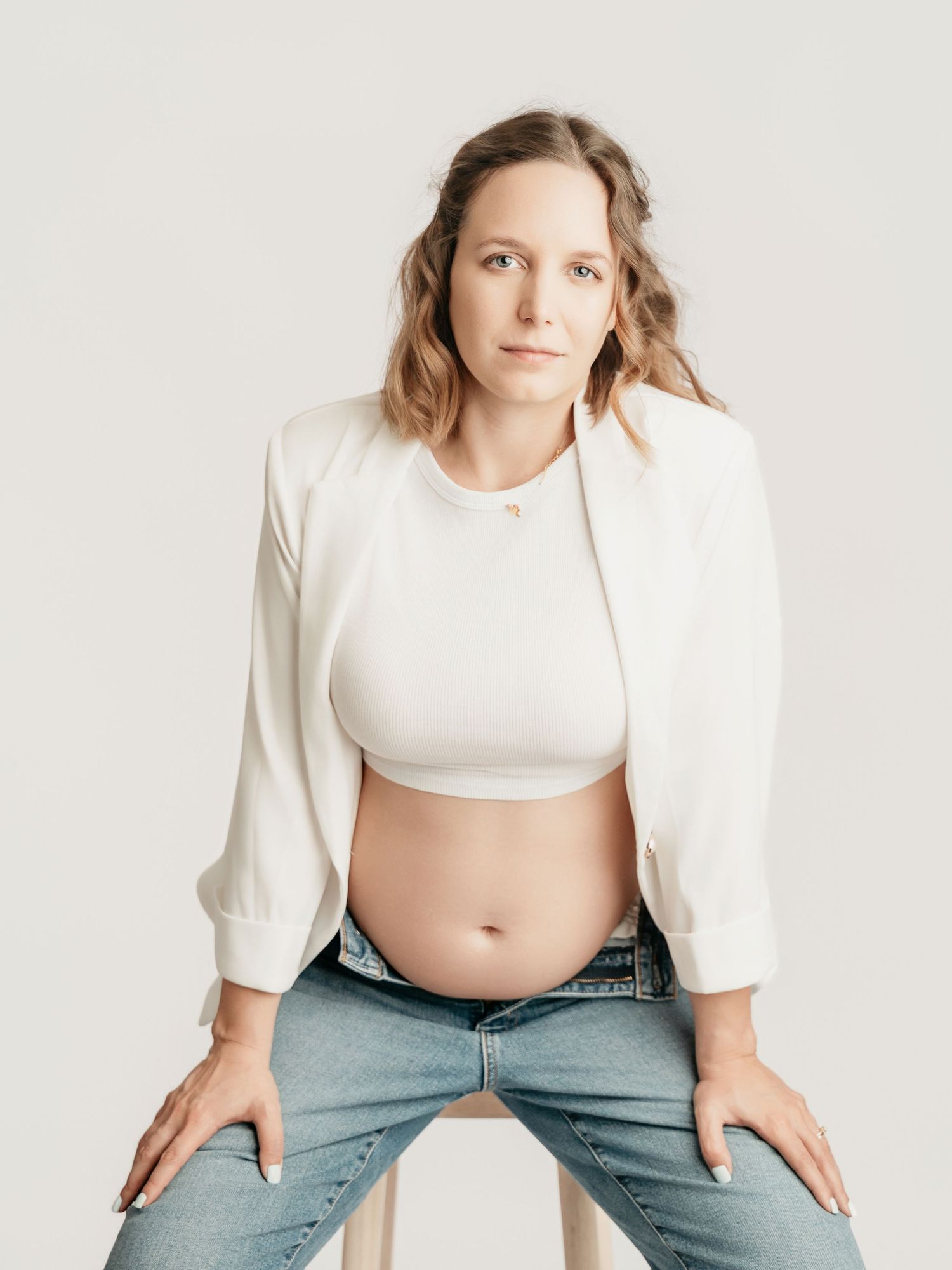 Pregnant woman in white crop top, blazer, and jeans sitting on stool showing baby bump.