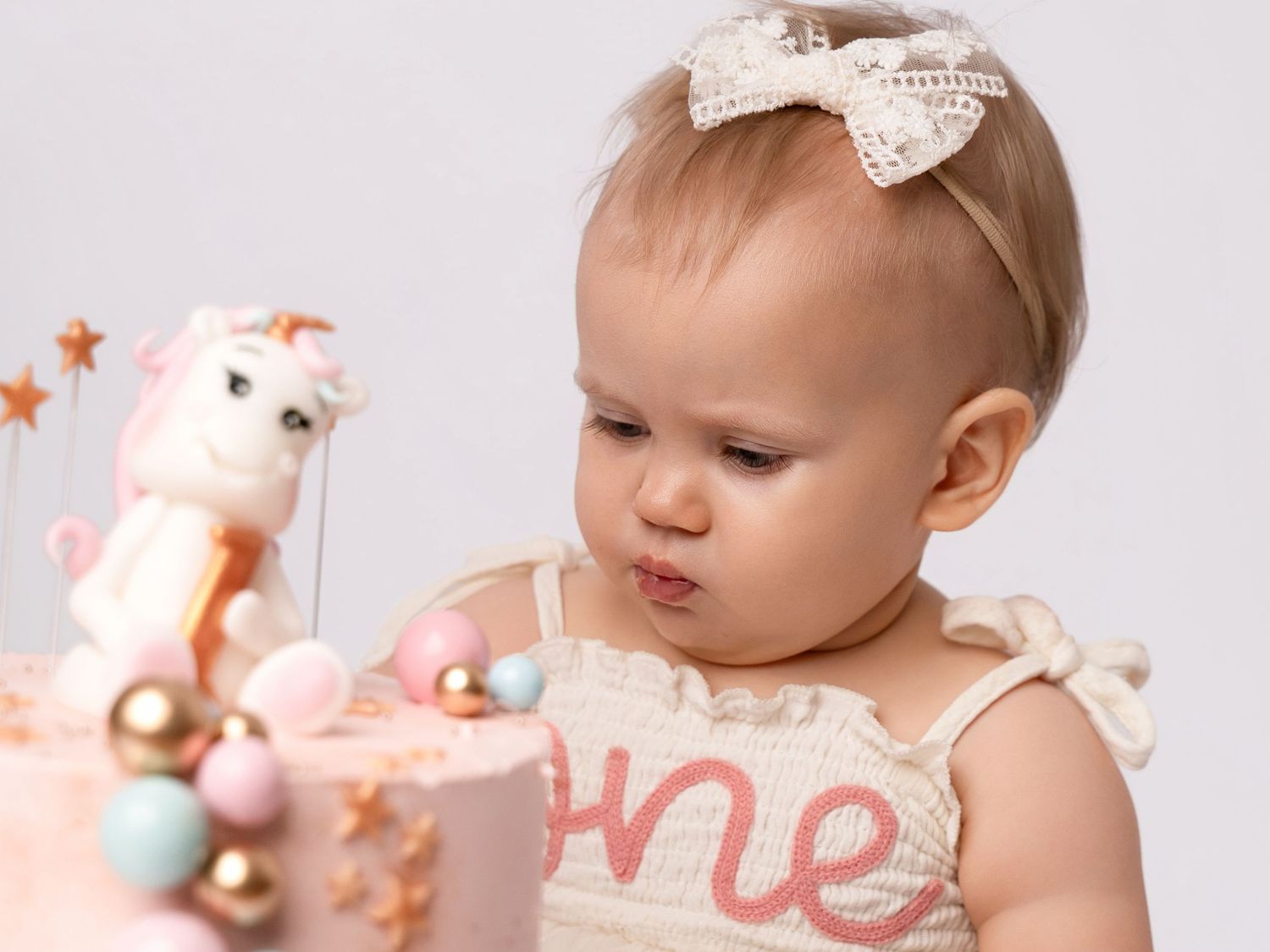 Baby girl in cream dress examines her unicorn first birthday cake with ONE topper.