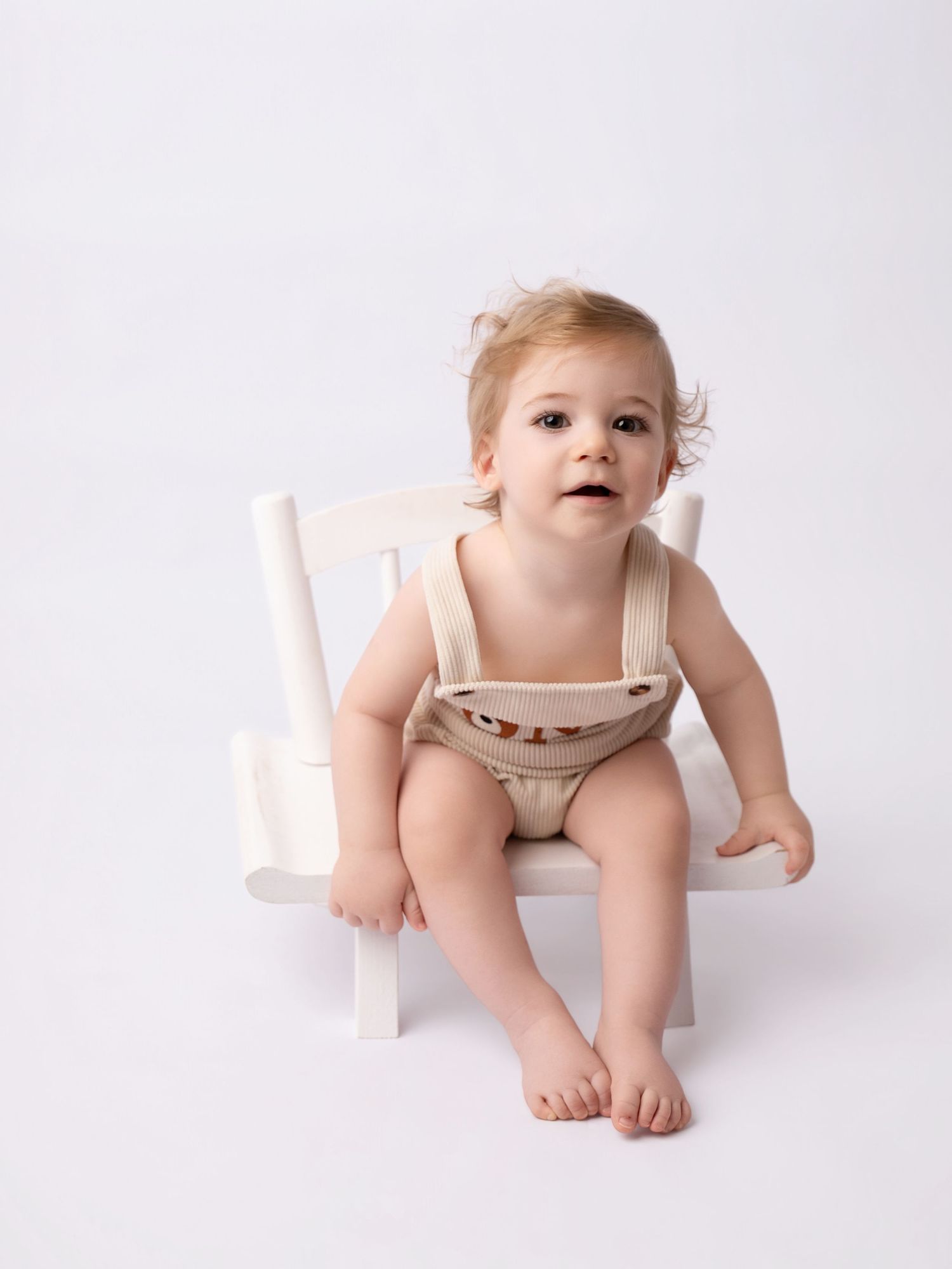 Adorable baby in beige overalls sitting on a small white chair against a white background.