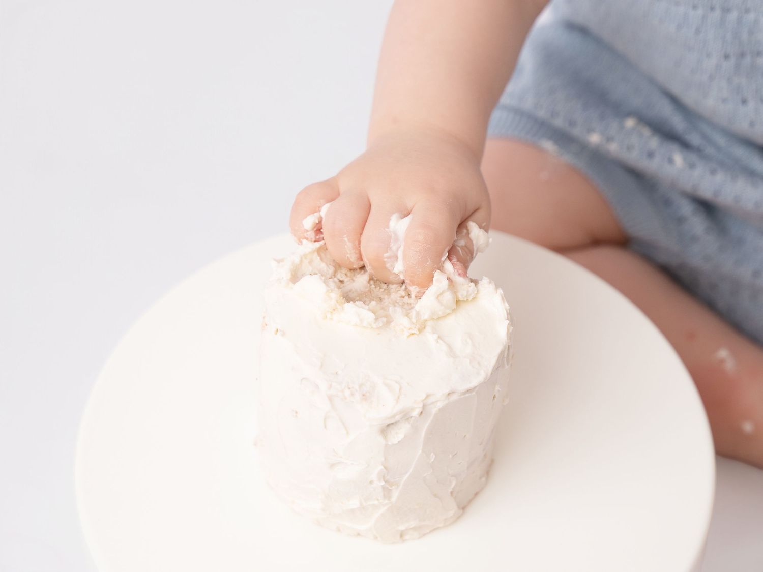 Baby hand smashing into a white frosted cake on a white pedestal during a first birthday cake smash photo session.