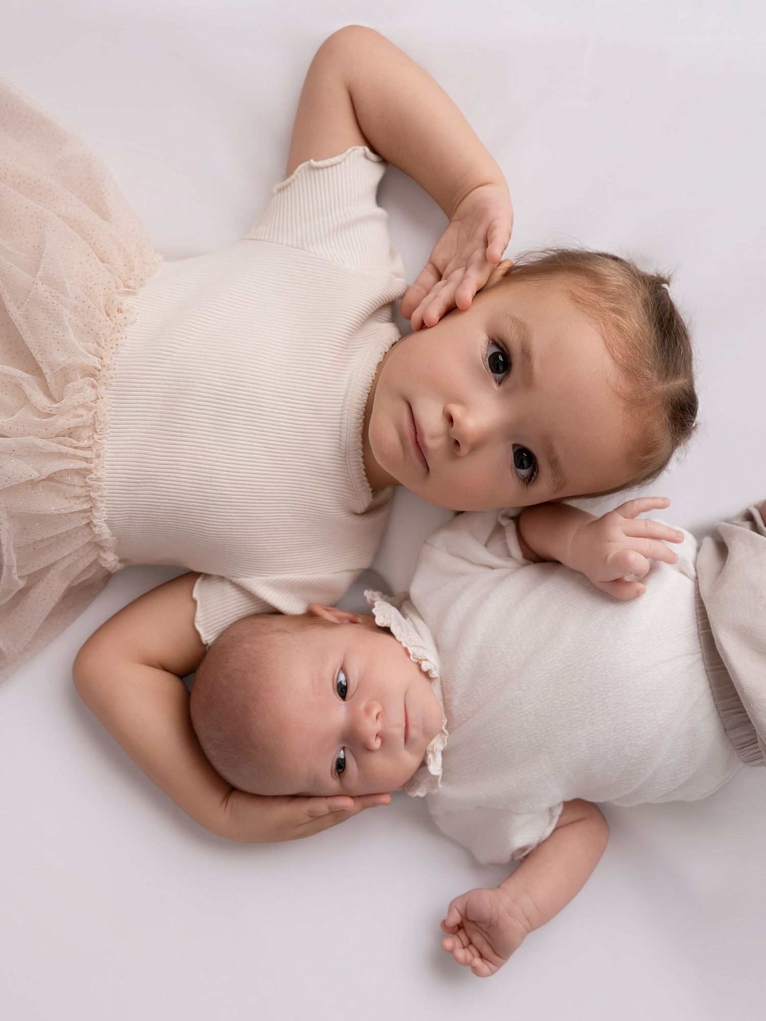 Two young siblings, a toddler and newborn baby, lying together in white outfits on a white background.