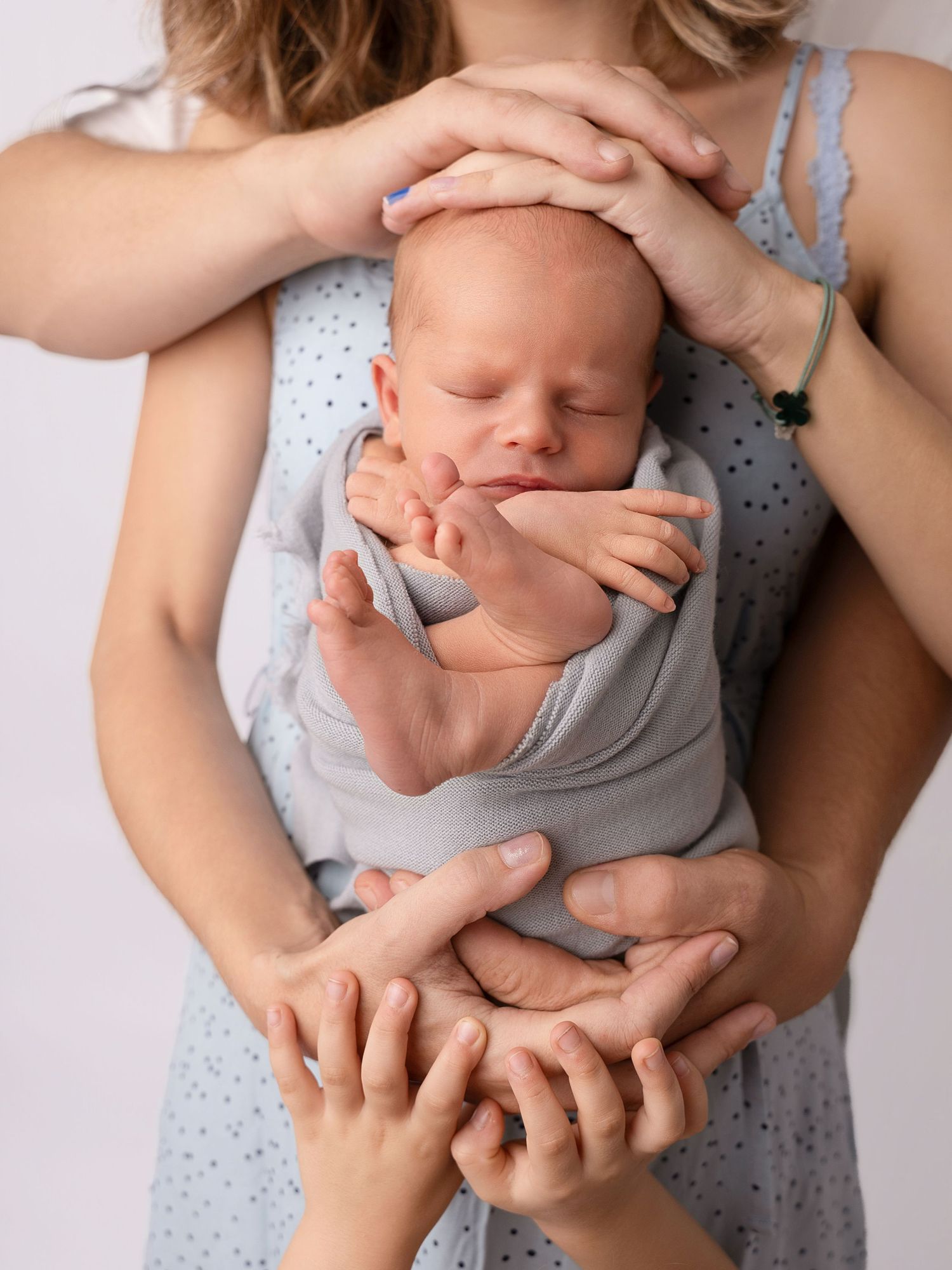 Newborn baby sleeping peacefully wrapped in gray, cradled by multiple loving hands against a white background.