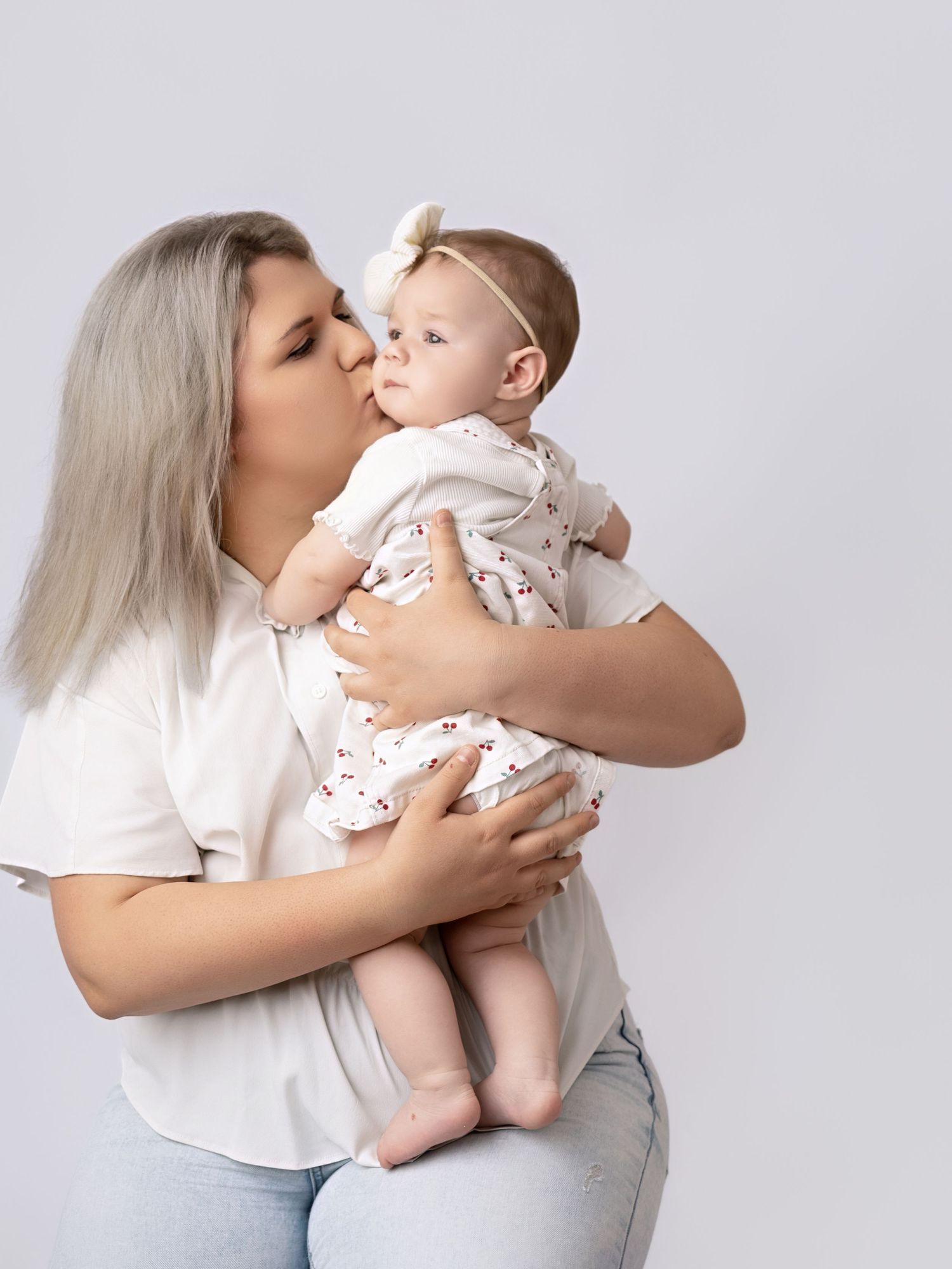 A mother with blonde hair kisses her baby girl in a white onesie and bow headband against a gray backdrop.
