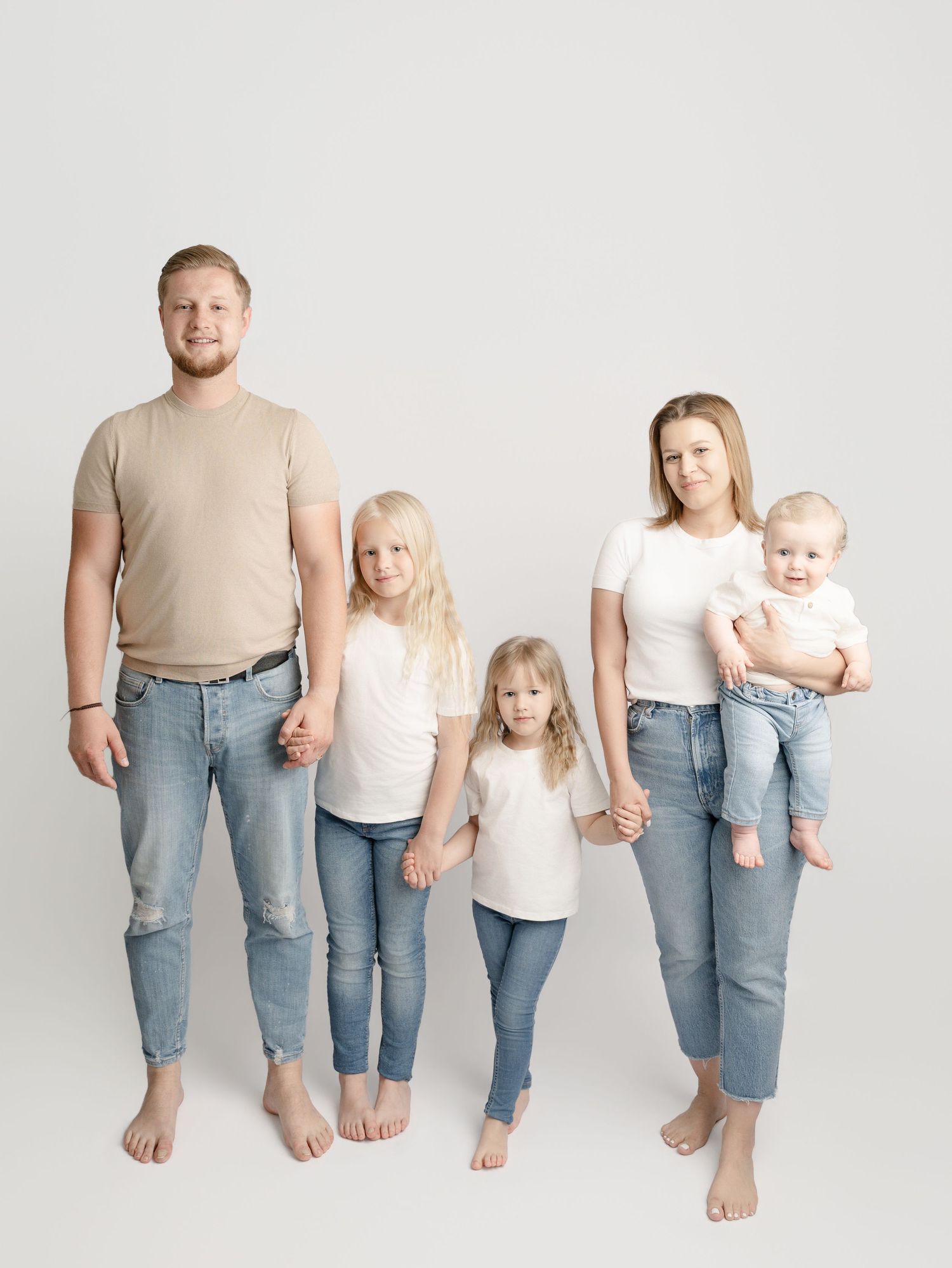 A family of five in matching casual outfits holds hands while posing barefoot in a studio portrait.