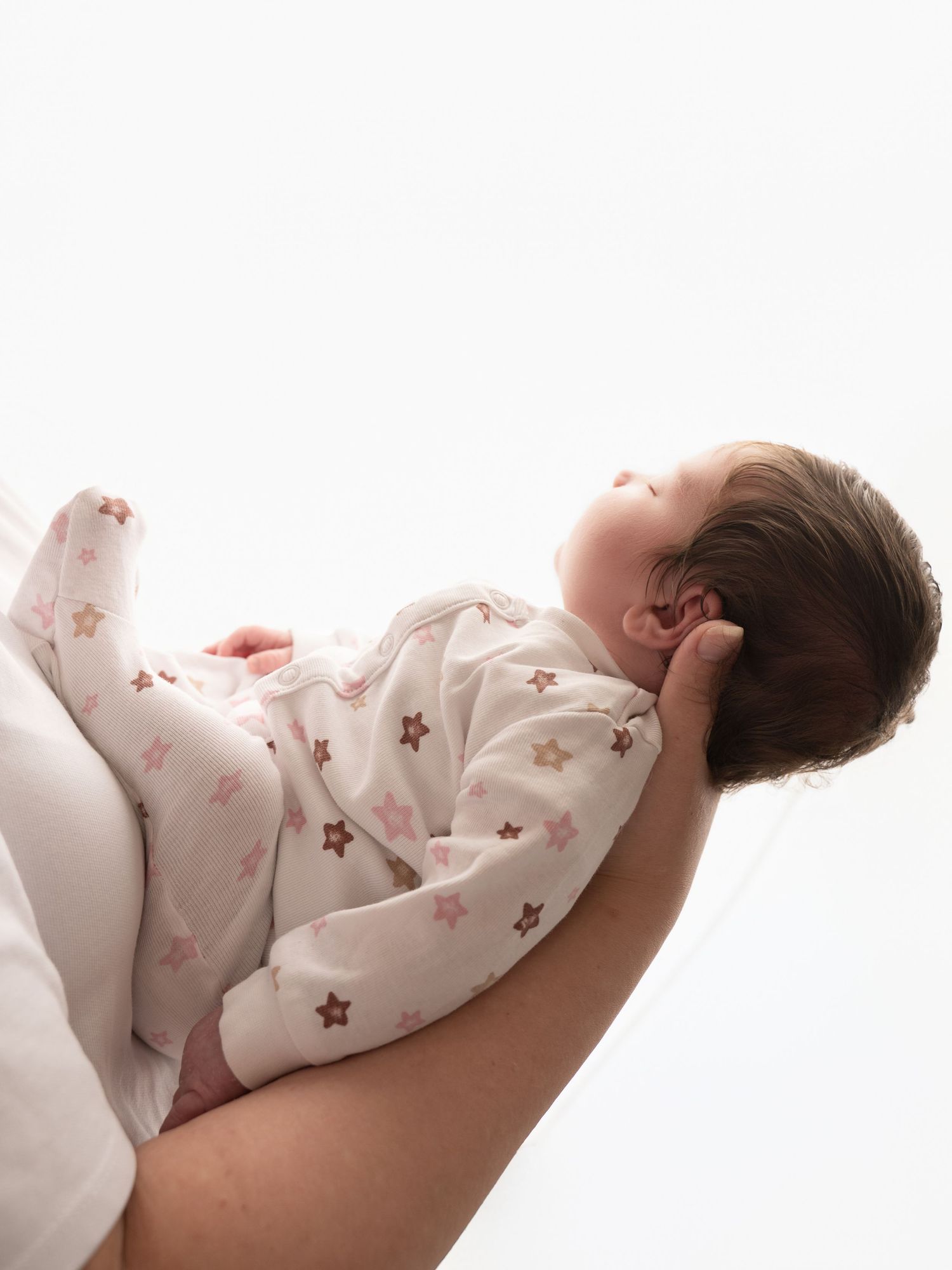 Sleeping newborn in star-print footie pajamas resting on mothers arm against bright white background.