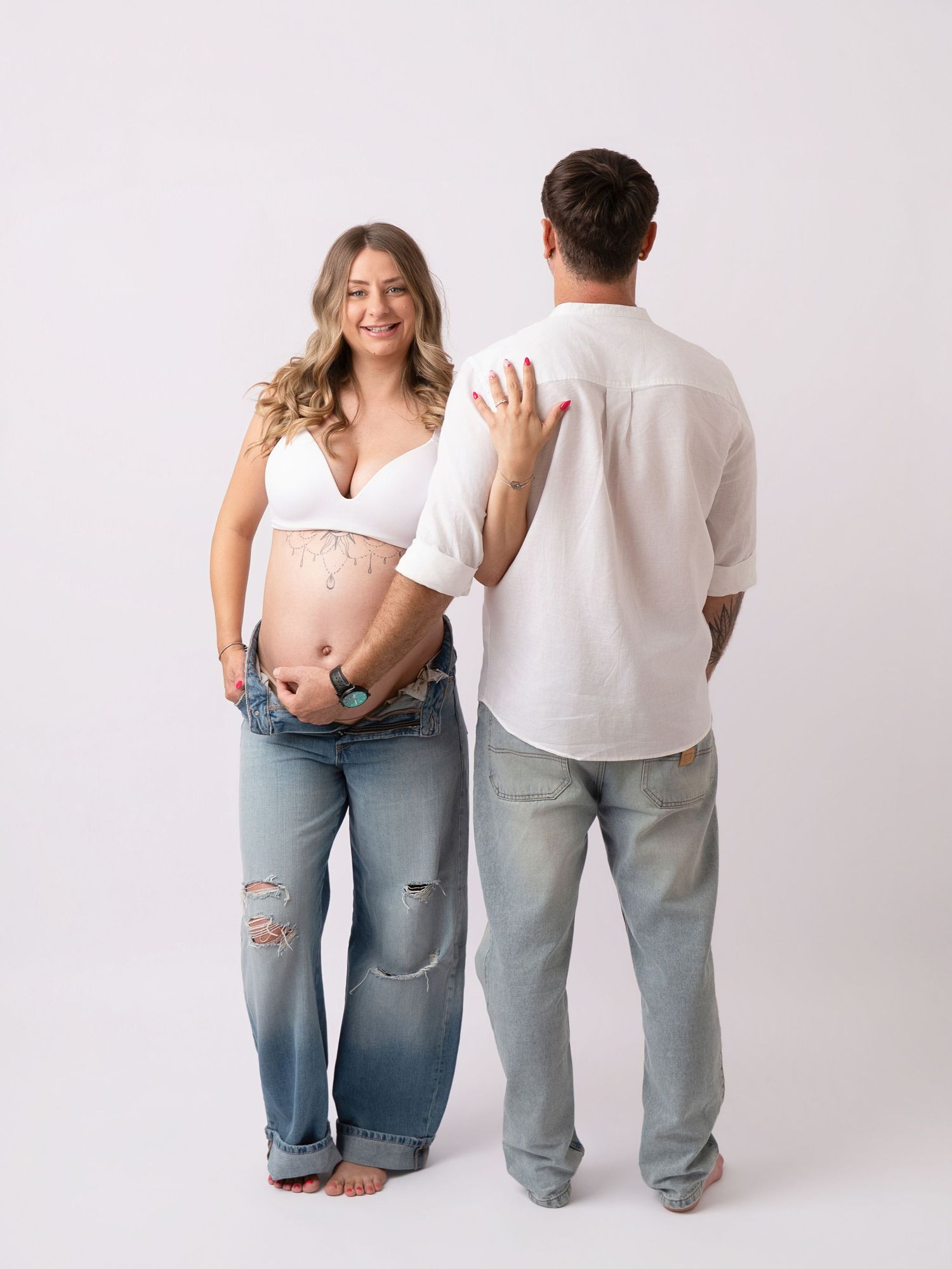 Pregnant woman in white crop top and jeans smiles beside man in white shirt during maternity photoshoot.