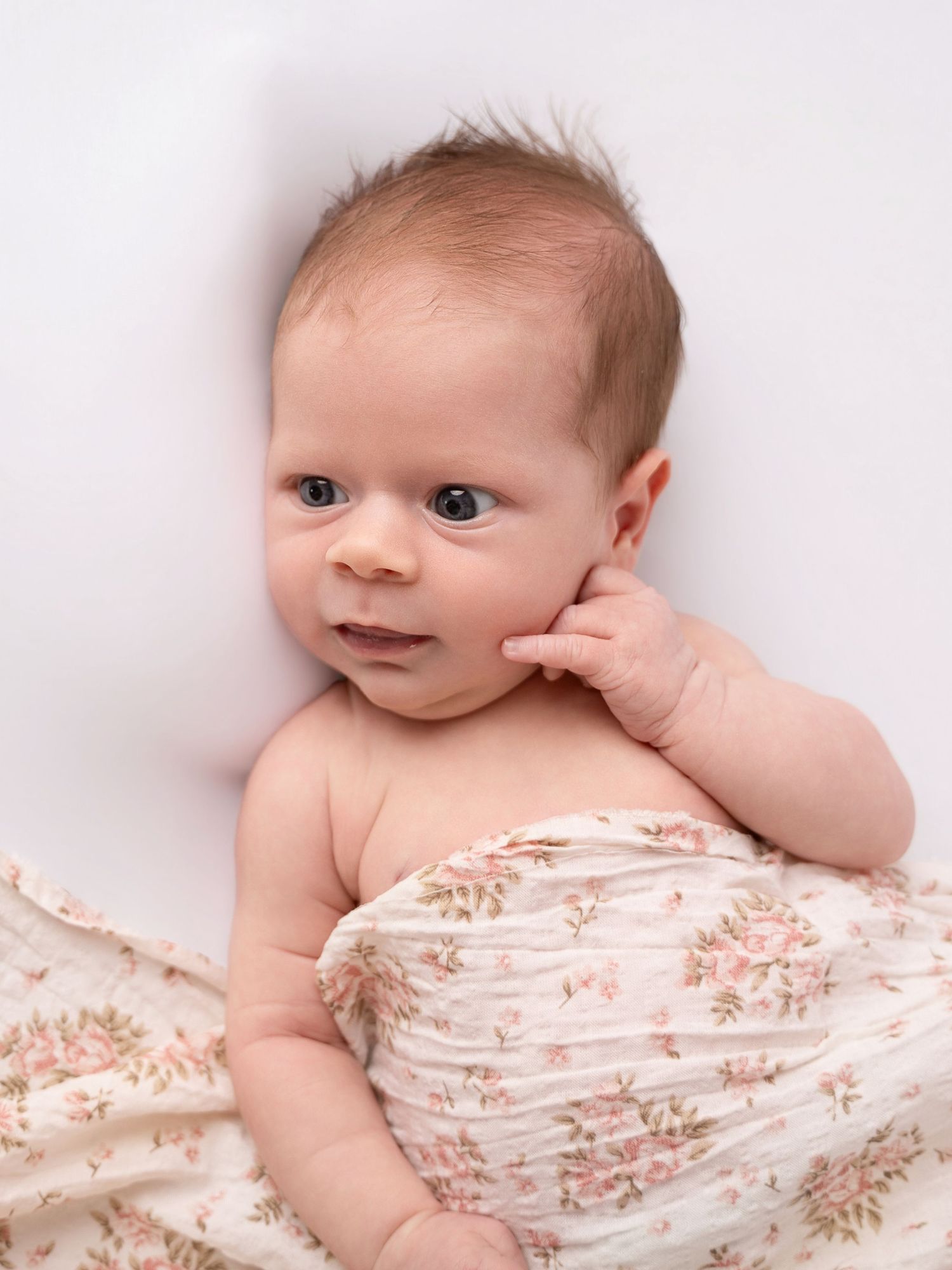 Awake newborn baby with dark hair wrapped in a floral muslin swaddle blanket lying on a white background.