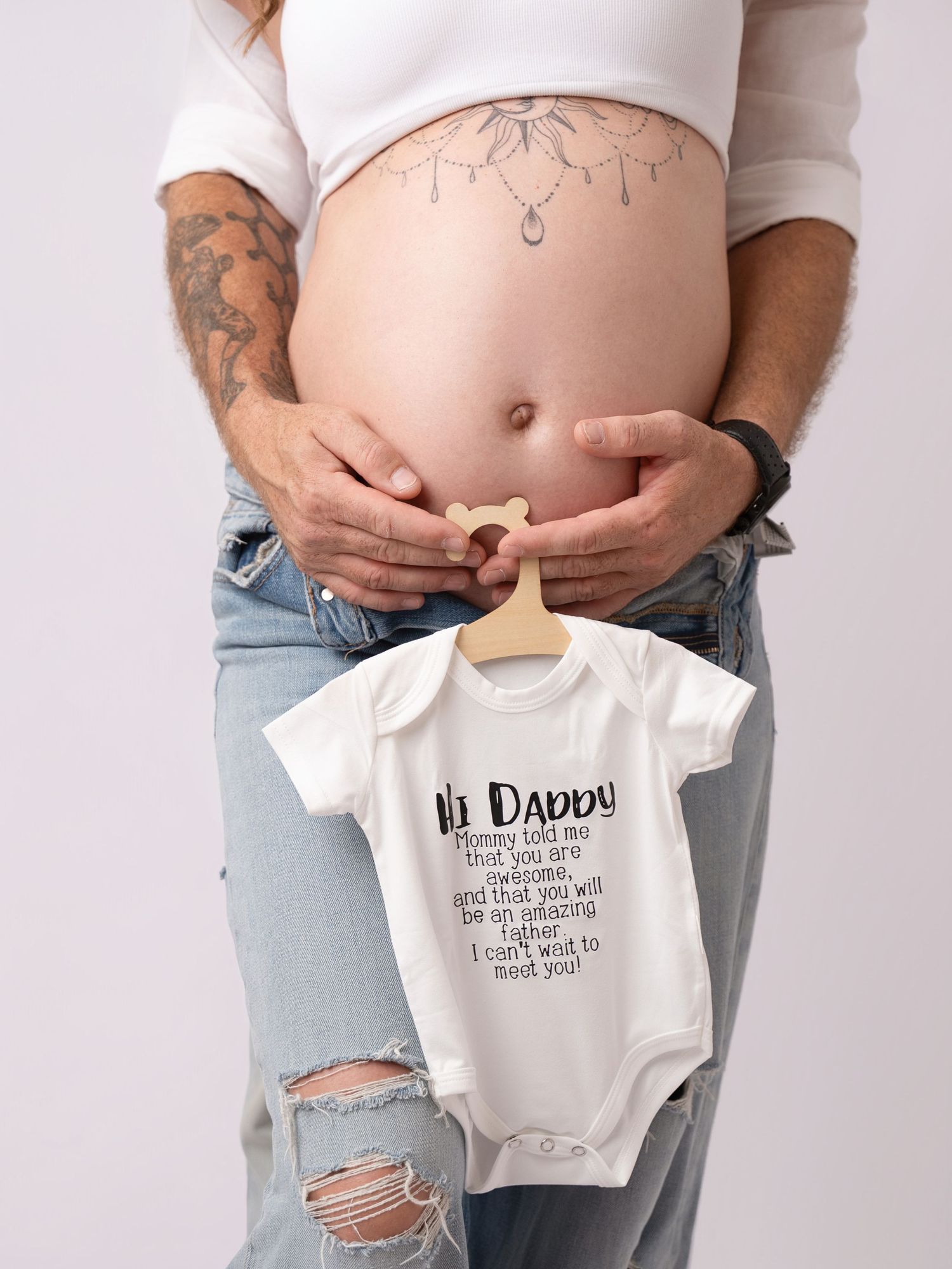 Pregnant woman holding a white baby onesie that reads Hi Daddy, I can't wait to meet you.