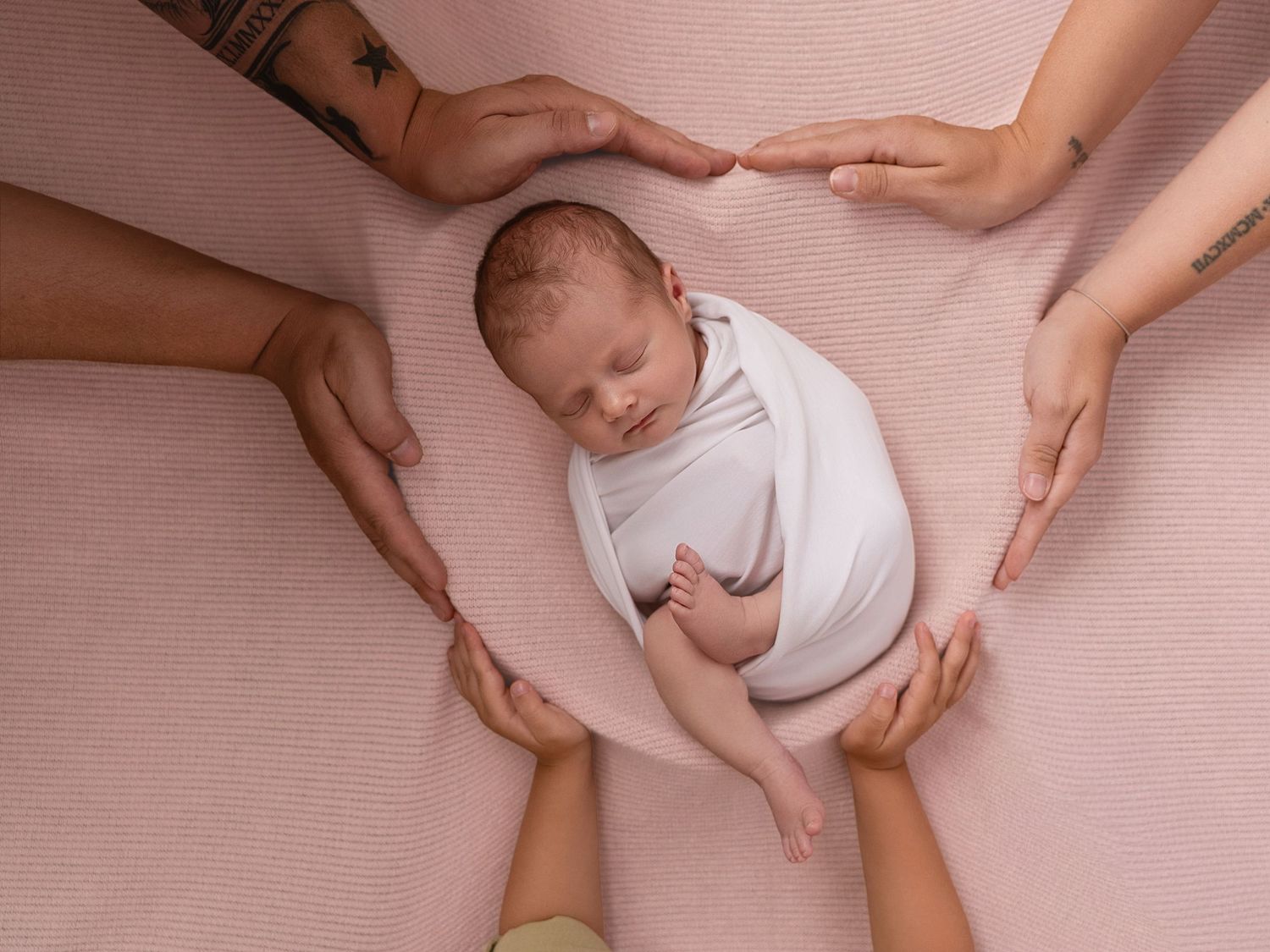 Newborn baby swaddled in white surrounded by multiple hands on a pink background.