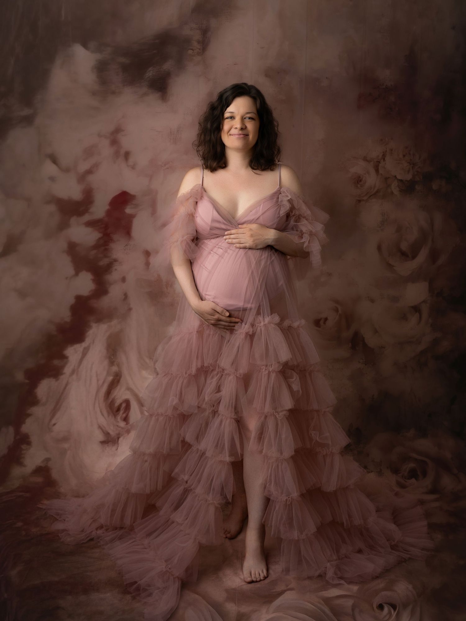 Smiling pregnant woman in ruffled pink tulle gown holding baby bump in elegant maternity portrait.