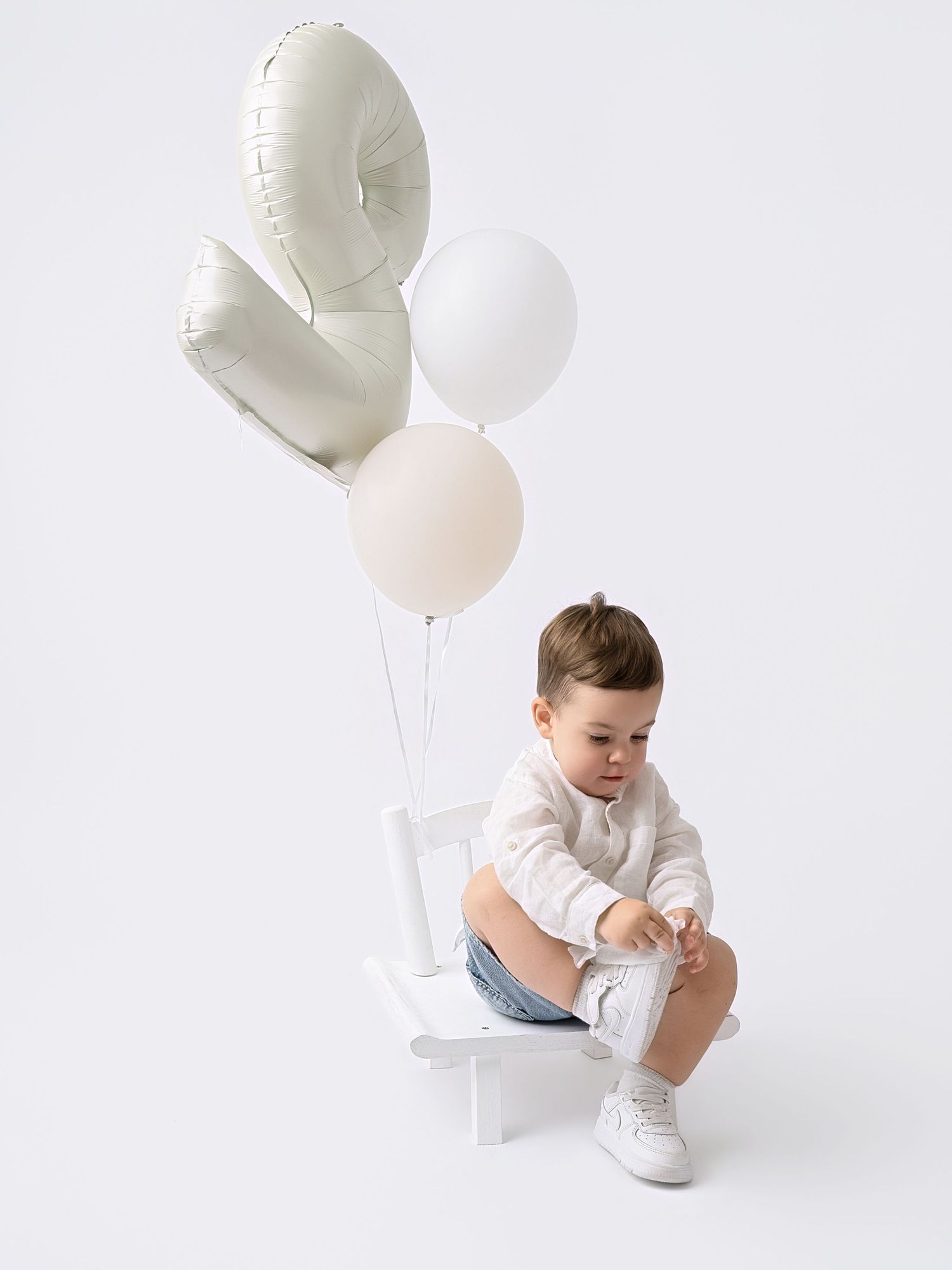 Toddler boy in white shirt and denim shorts sitting on small chair with white and beige balloons.