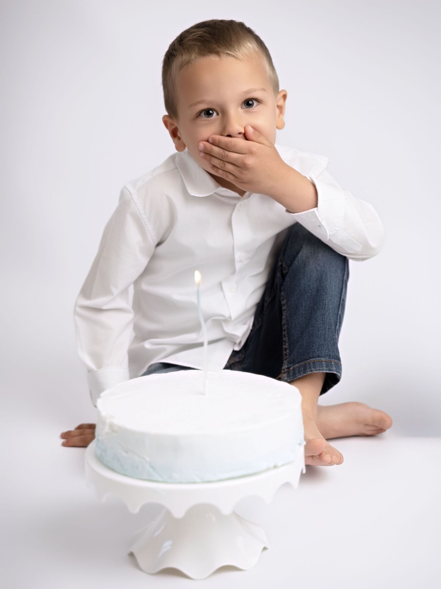 Young boy in white shirt sits cross-legged beside a blue birthday cake with one candle, hand covering his mouth.