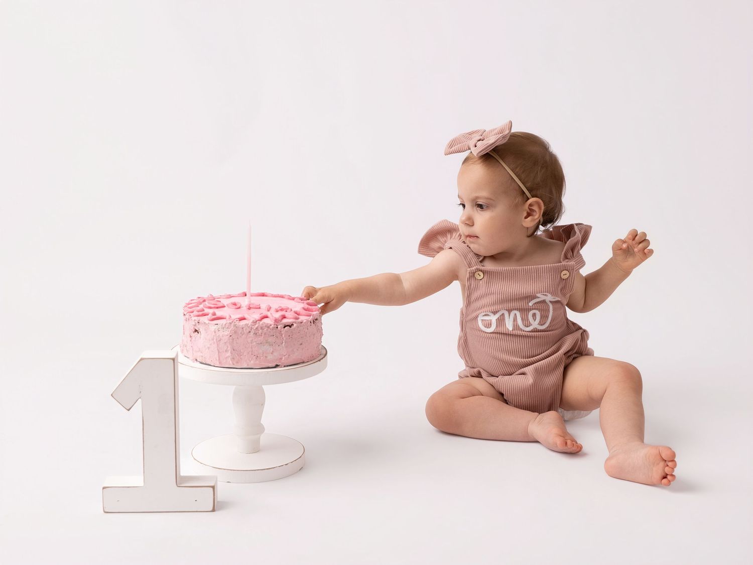 Baby girl in pink romper touching her first birthday cake next to a number one decoration on white background.