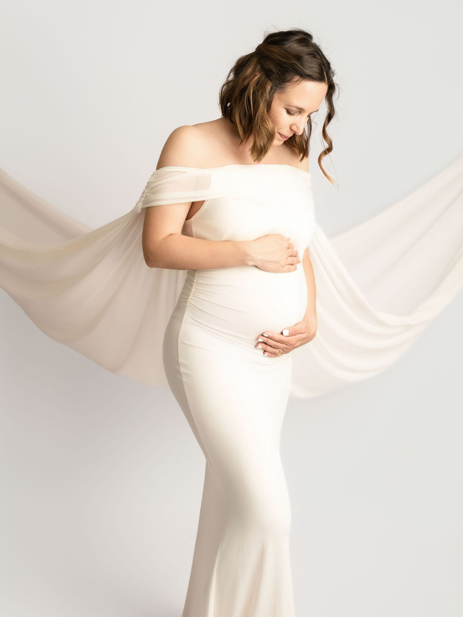 Pregnant woman in fitted ivory off-shoulder maternity dress with flowing chiffon wrap in studio.