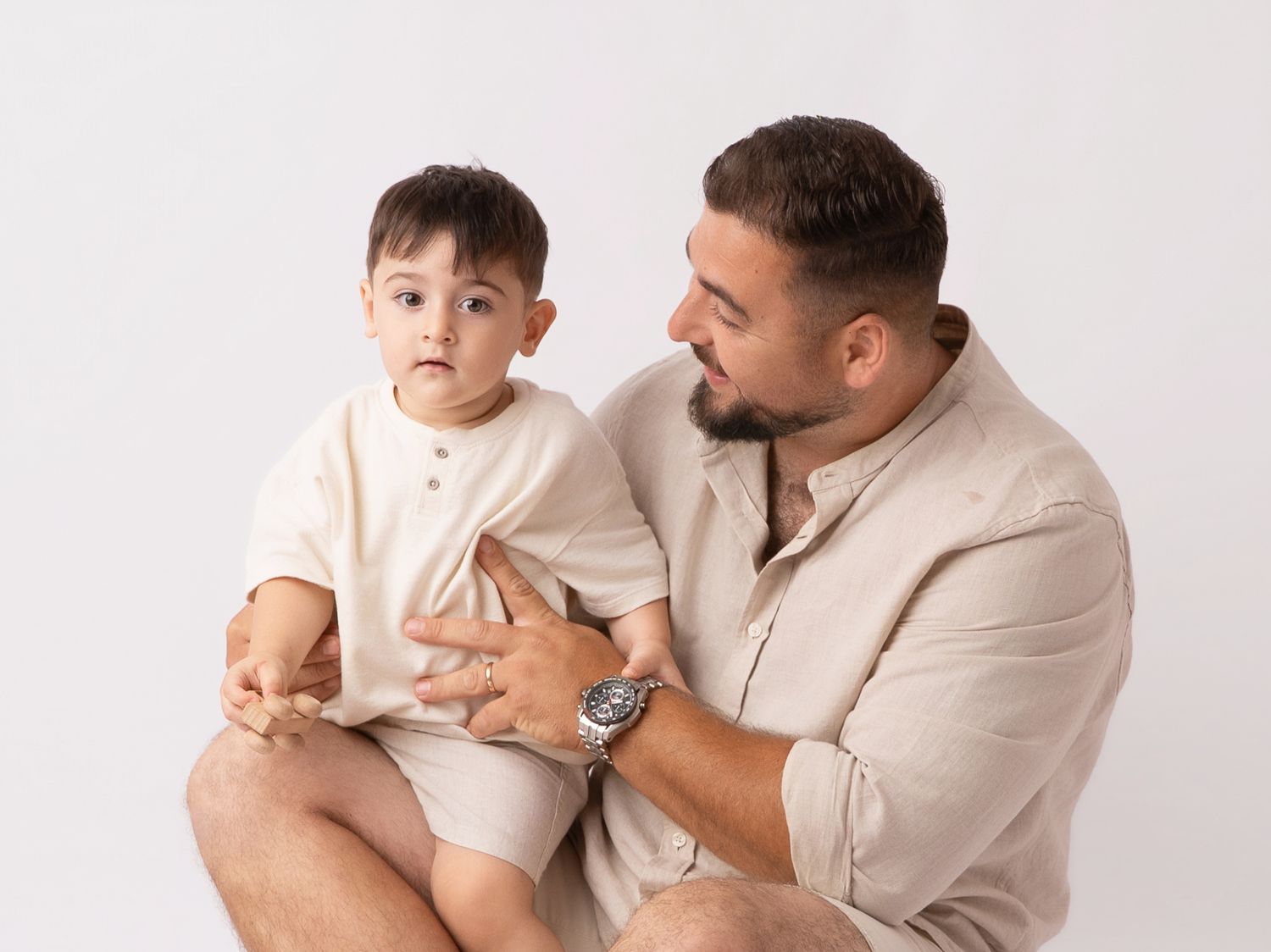 A father holds his toddler son on his lap during a professional studio portrait session on white background.