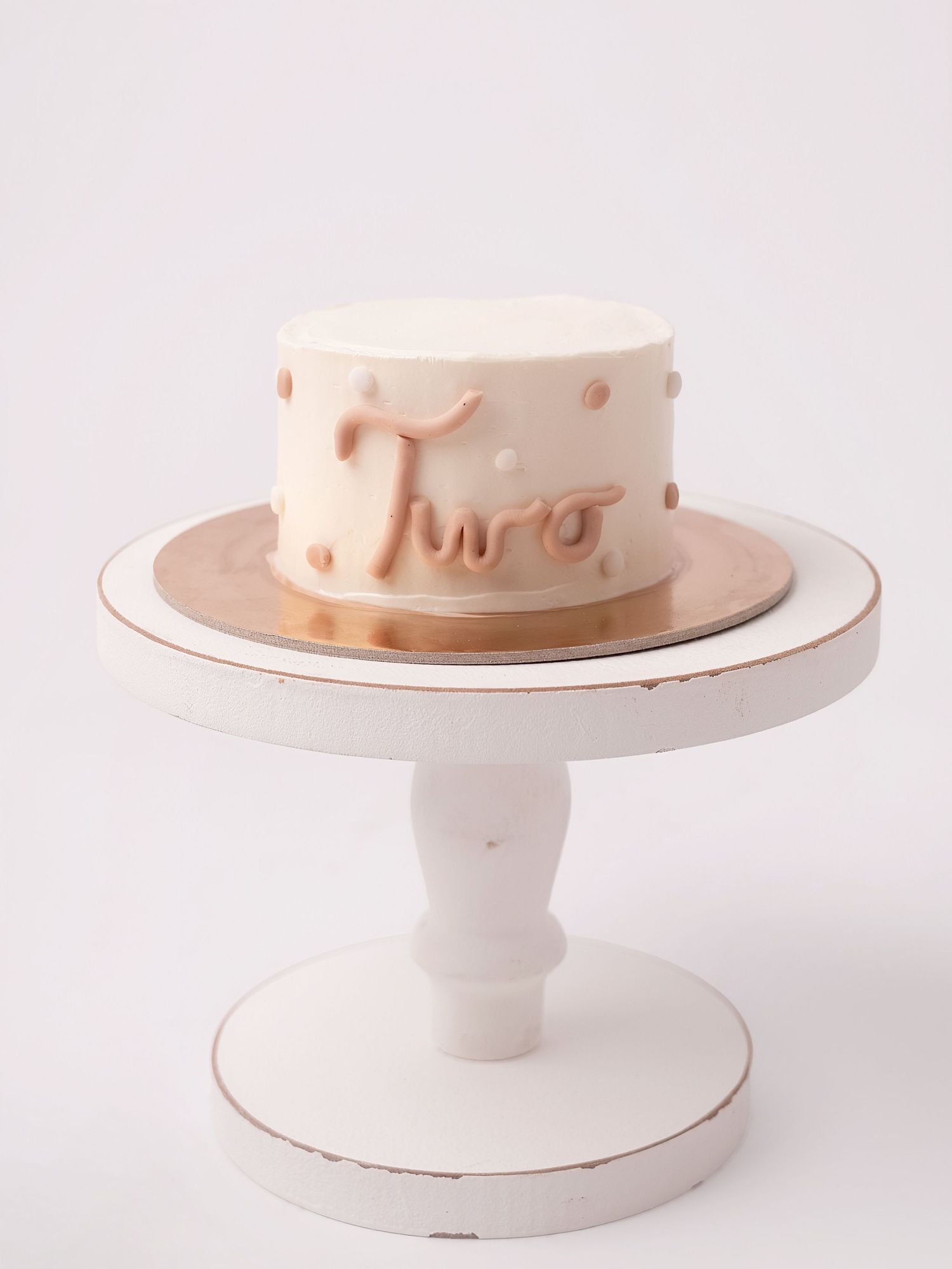 White and rose gold 'Two' birthday cake with polka dots on a distressed white pedestal cake stand.