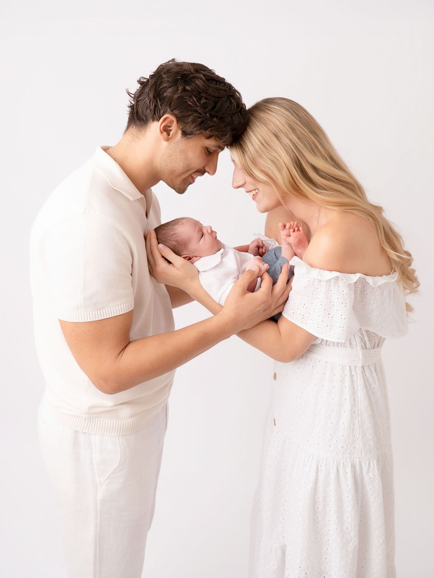 A couple in white outfits tenderly holds their newborn twins, foreheads touching, against a white background.