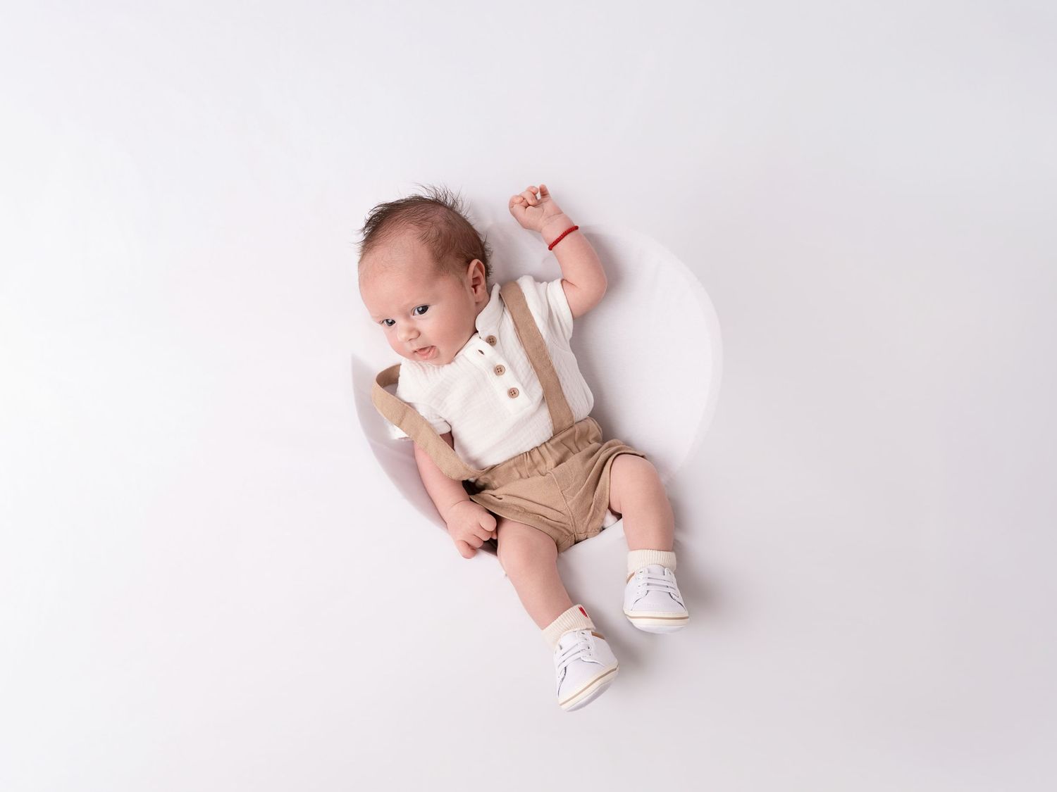 Newborn baby in cream suspender outfit and white shoes lying on white background with one fist raised.