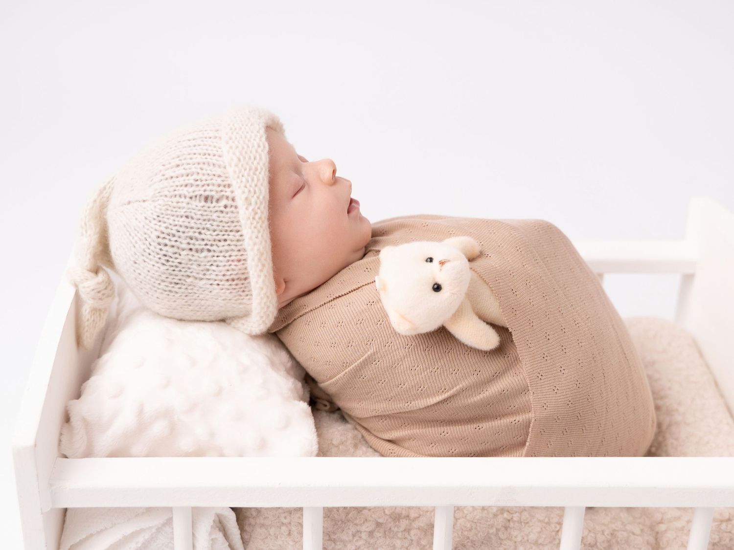 Sleeping newborn baby in knit hat holding teddy bear in white wooden crib on white background.