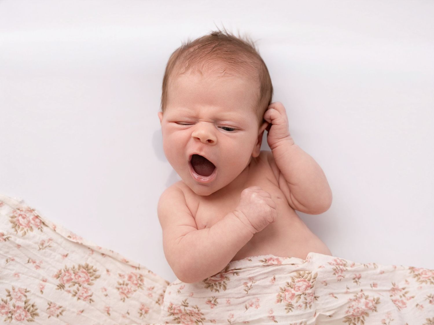 Newborn baby yawning while lying on a floral blanket against a white background.