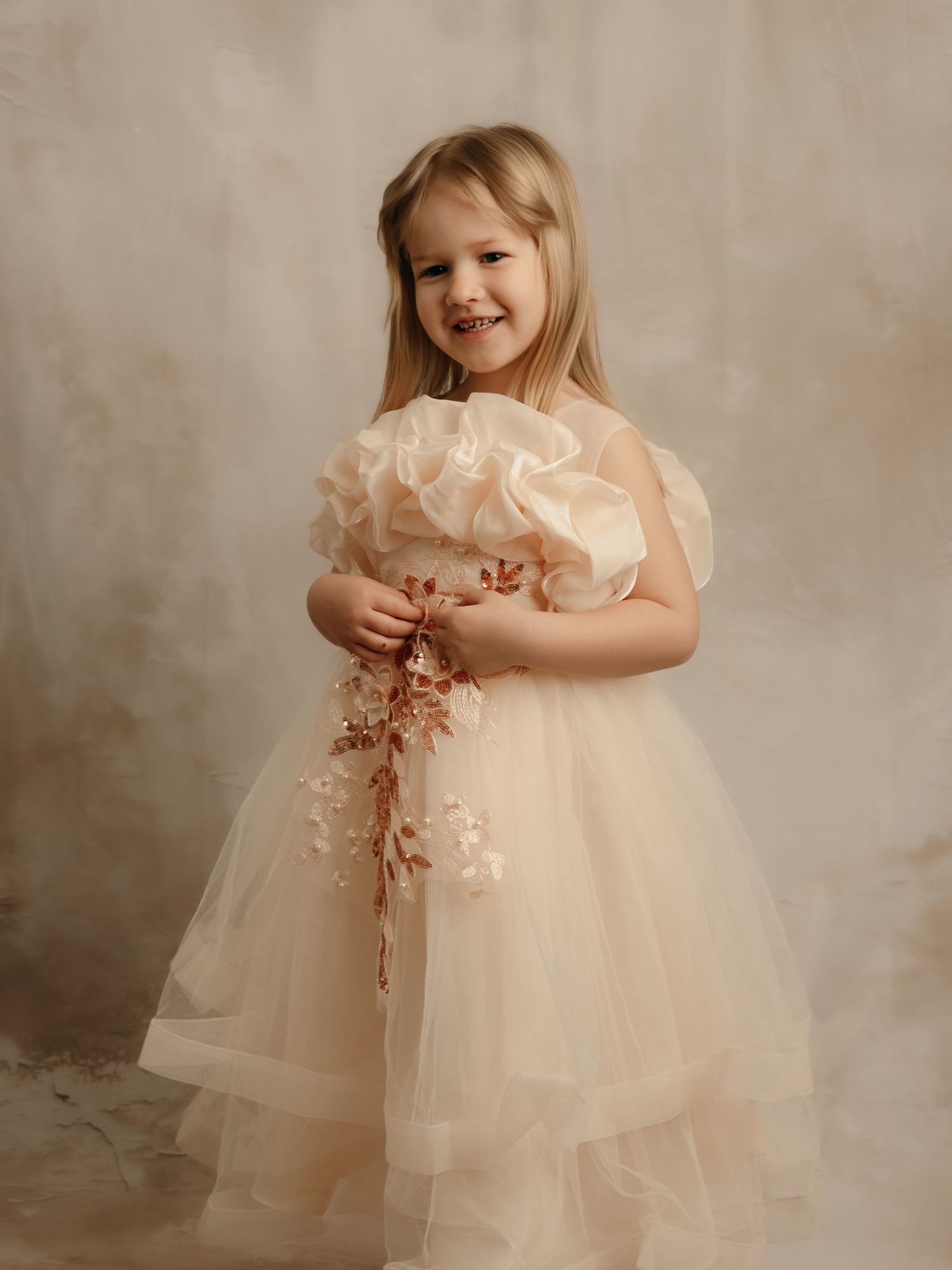 Happy girl in a champagne ruffled tulle dress holding a delicate floral branch in a studio portrait.