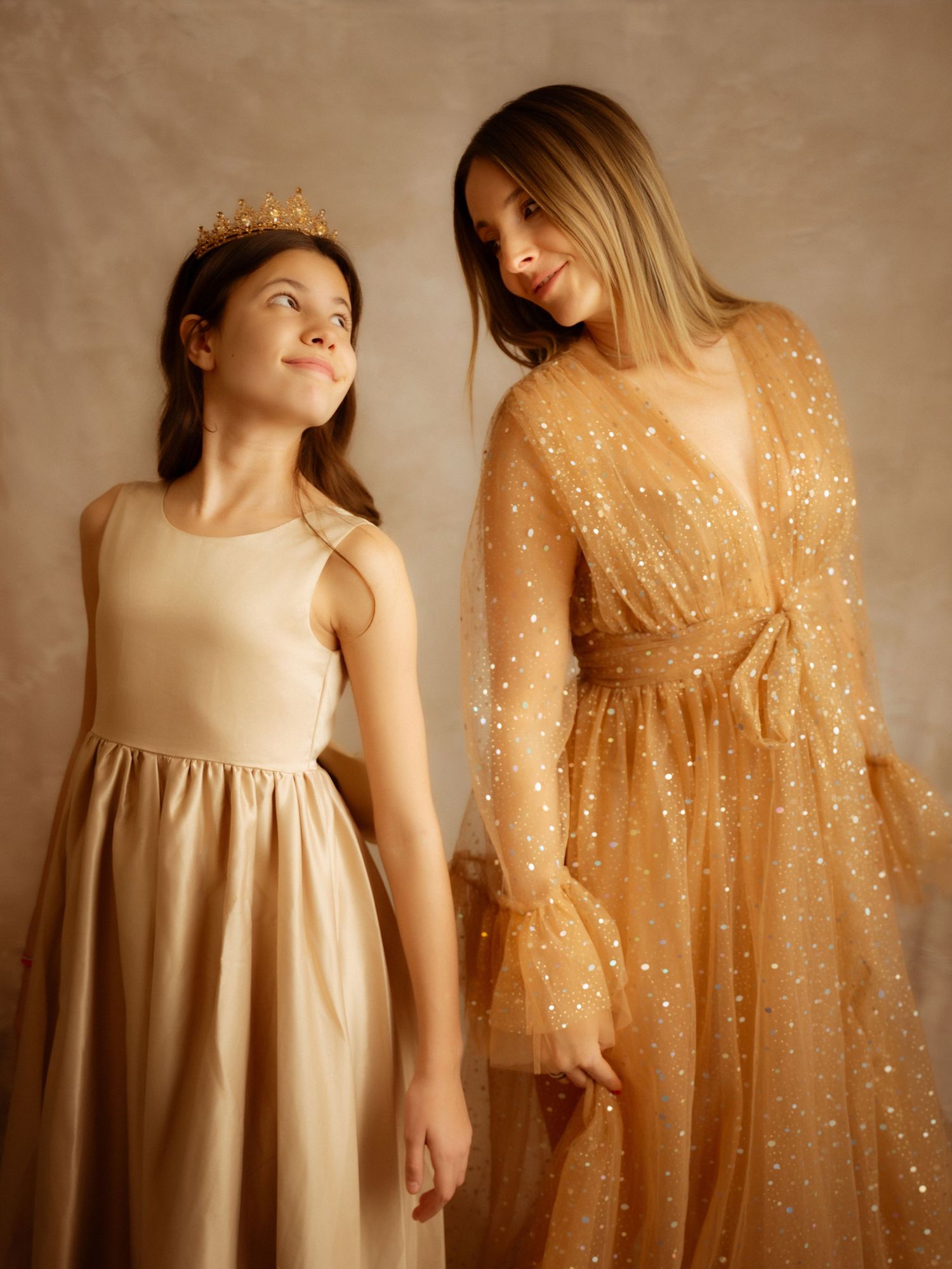 A mother in a sparkling golden gown and daughter in cream dress share a joyful studio portrait moment.