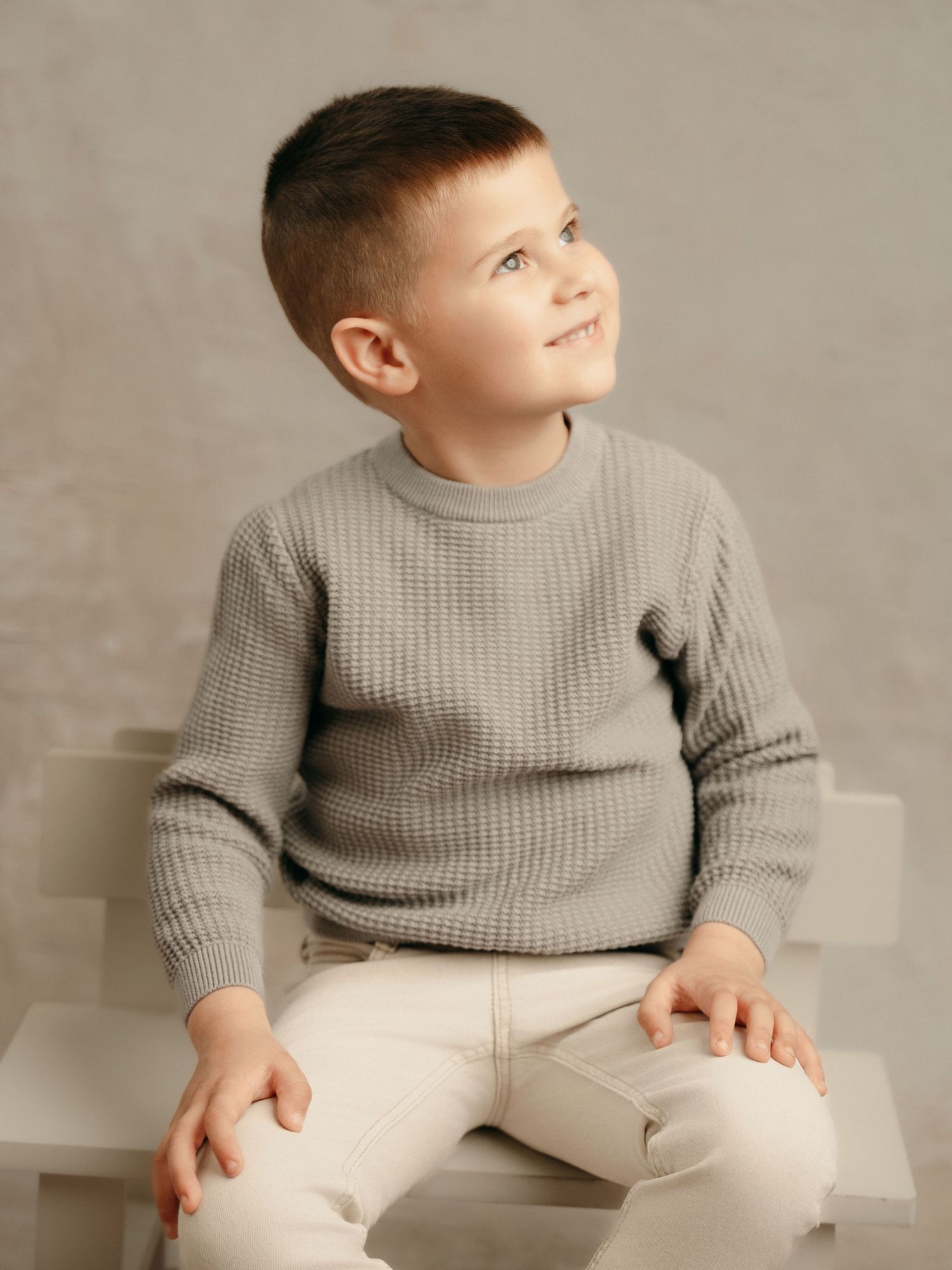 Cute young boy in gray knit sweater and beige pants sitting and smiling in a studio portrait.
