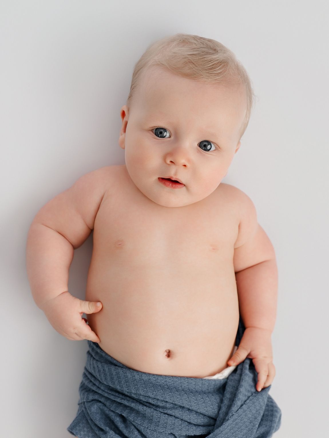 Adorable baby with blue eyes lying down, wrapped in a blue blanket against a white background.