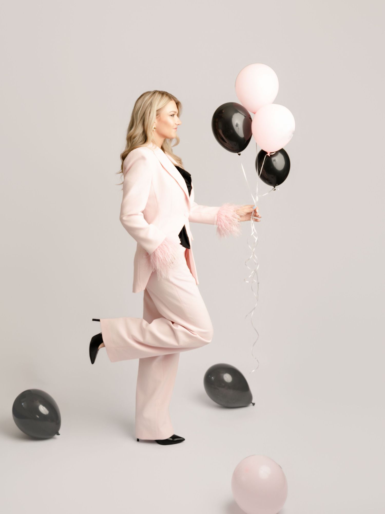 A blonde woman in a pink blazer suit holds pink and black balloons while posing playfully in a studio.