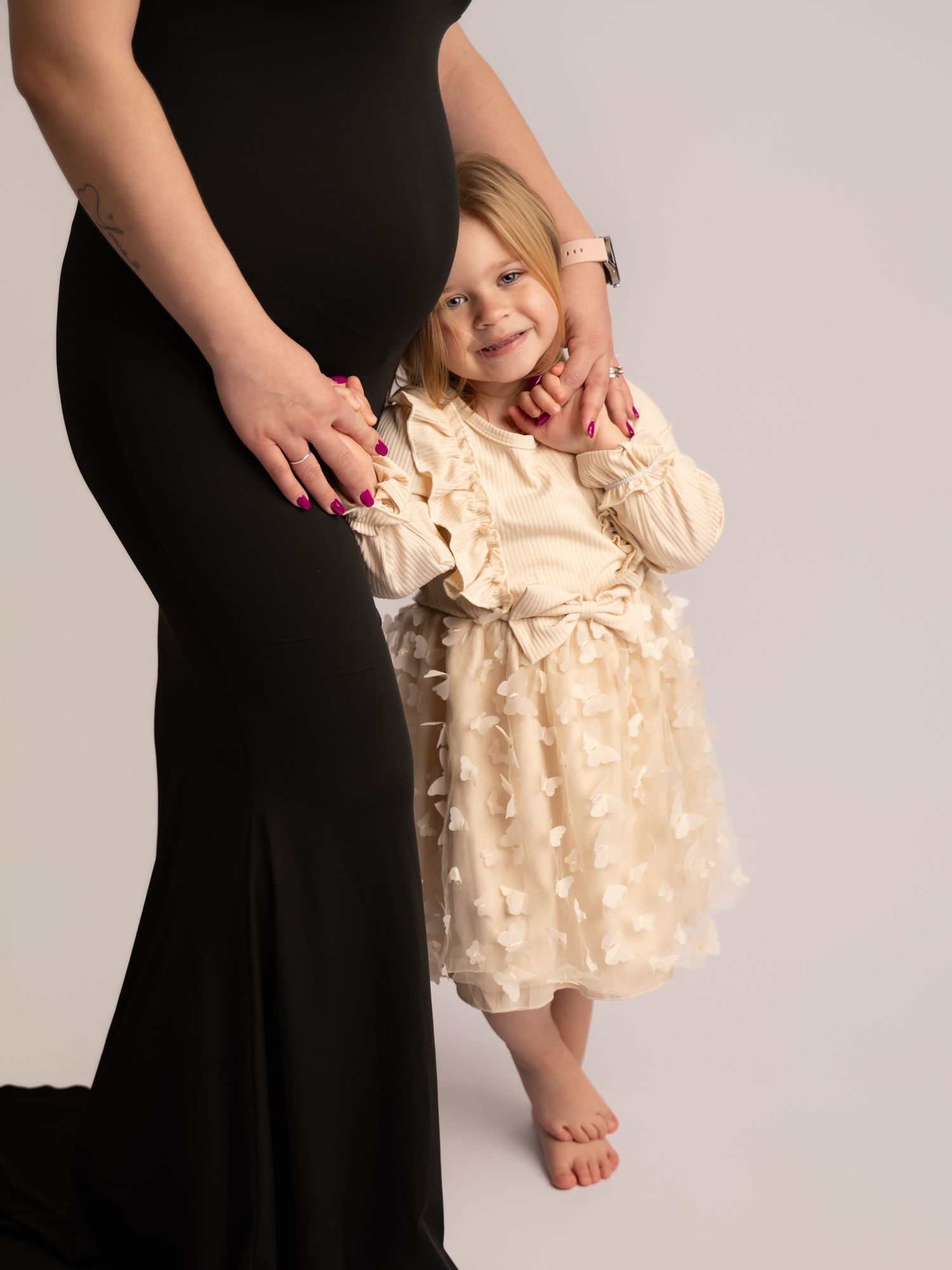 Smiling toddler girl in cream butterfly dress standing beside pregnant woman in black gown.