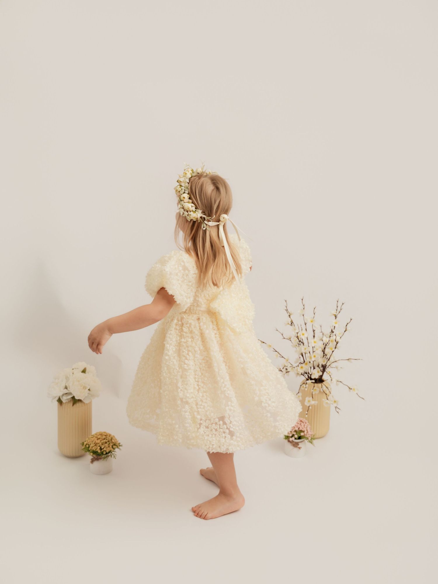 A young girl in a cream puff-sleeve dress with floral crown twirls among white and gold floral arrangements.