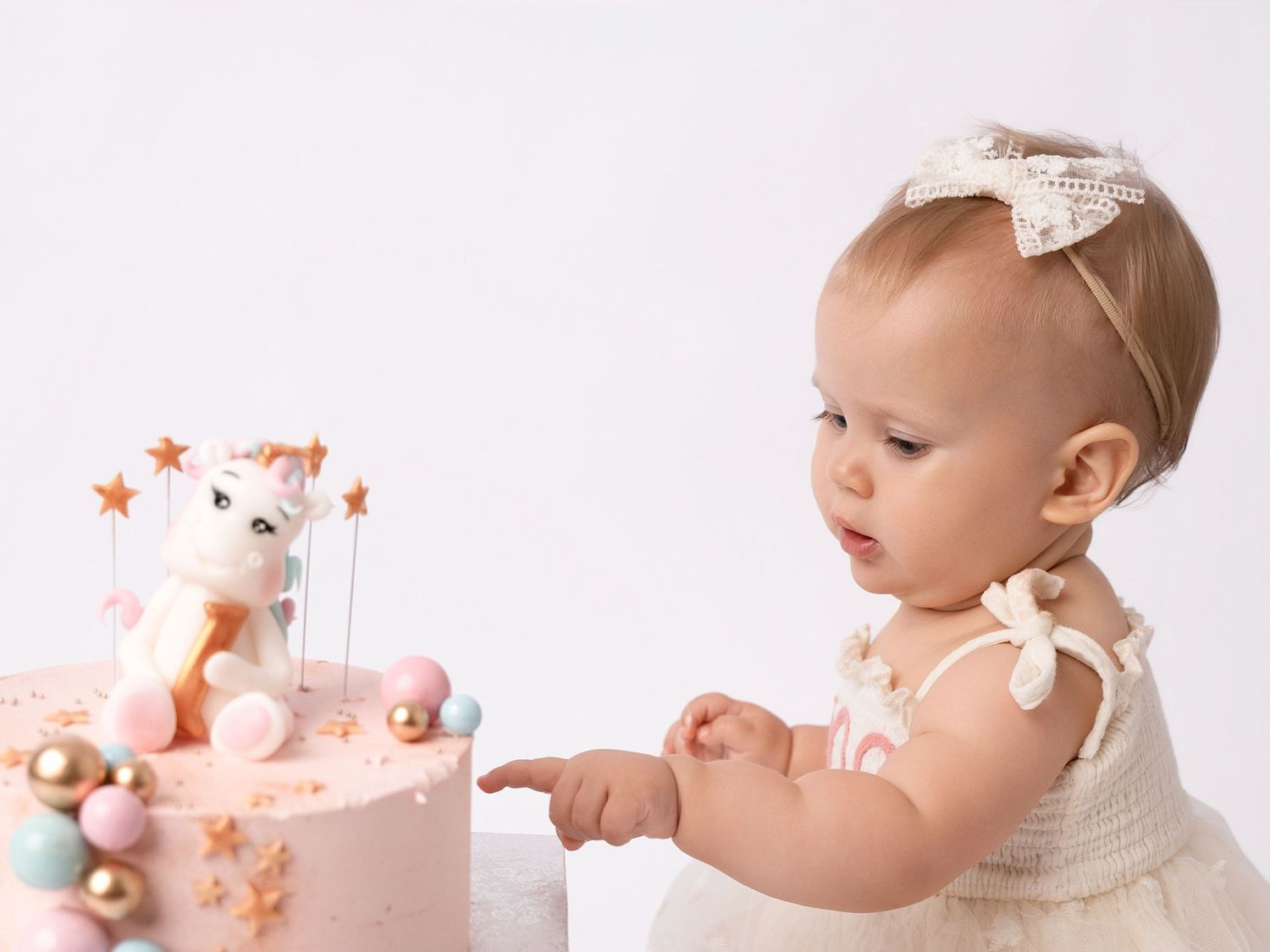 Baby girl in white dress pointing at a pink unicorn first birthday cake on a white background.