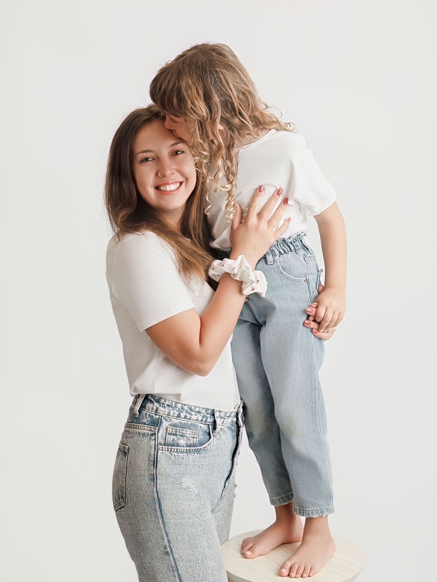 A smiling woman in white tee and jeans holds a young girl in matching outfit against a white background.