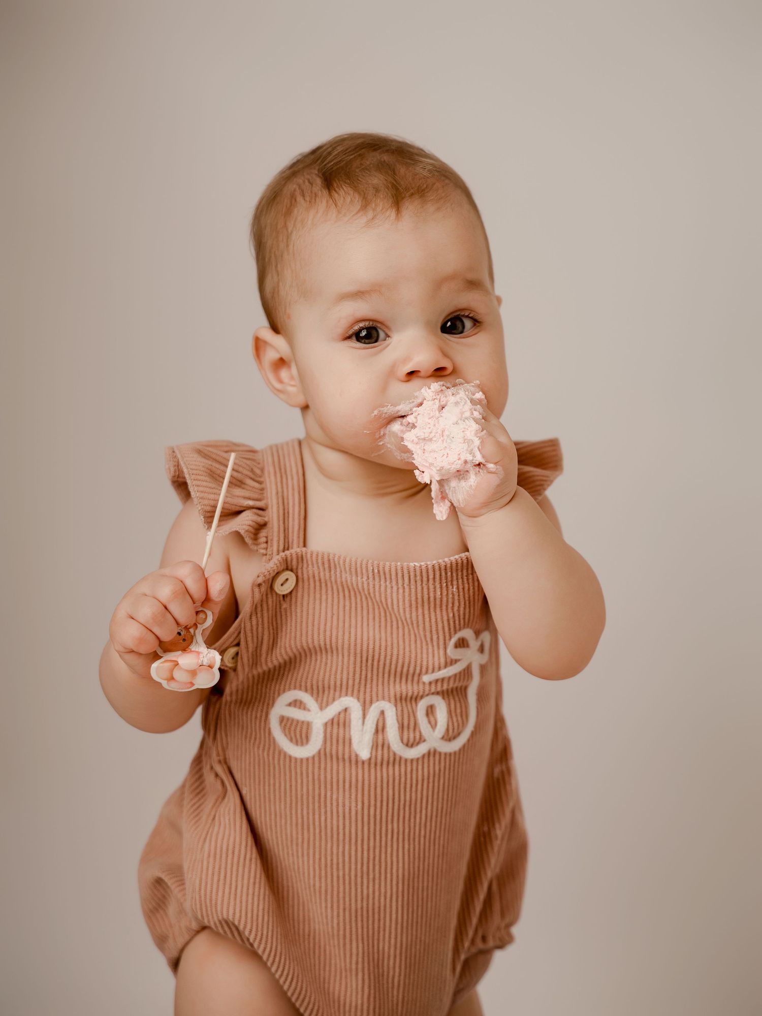 Baby in a brown 'one' romper eating smash cake during first birthday photo session.