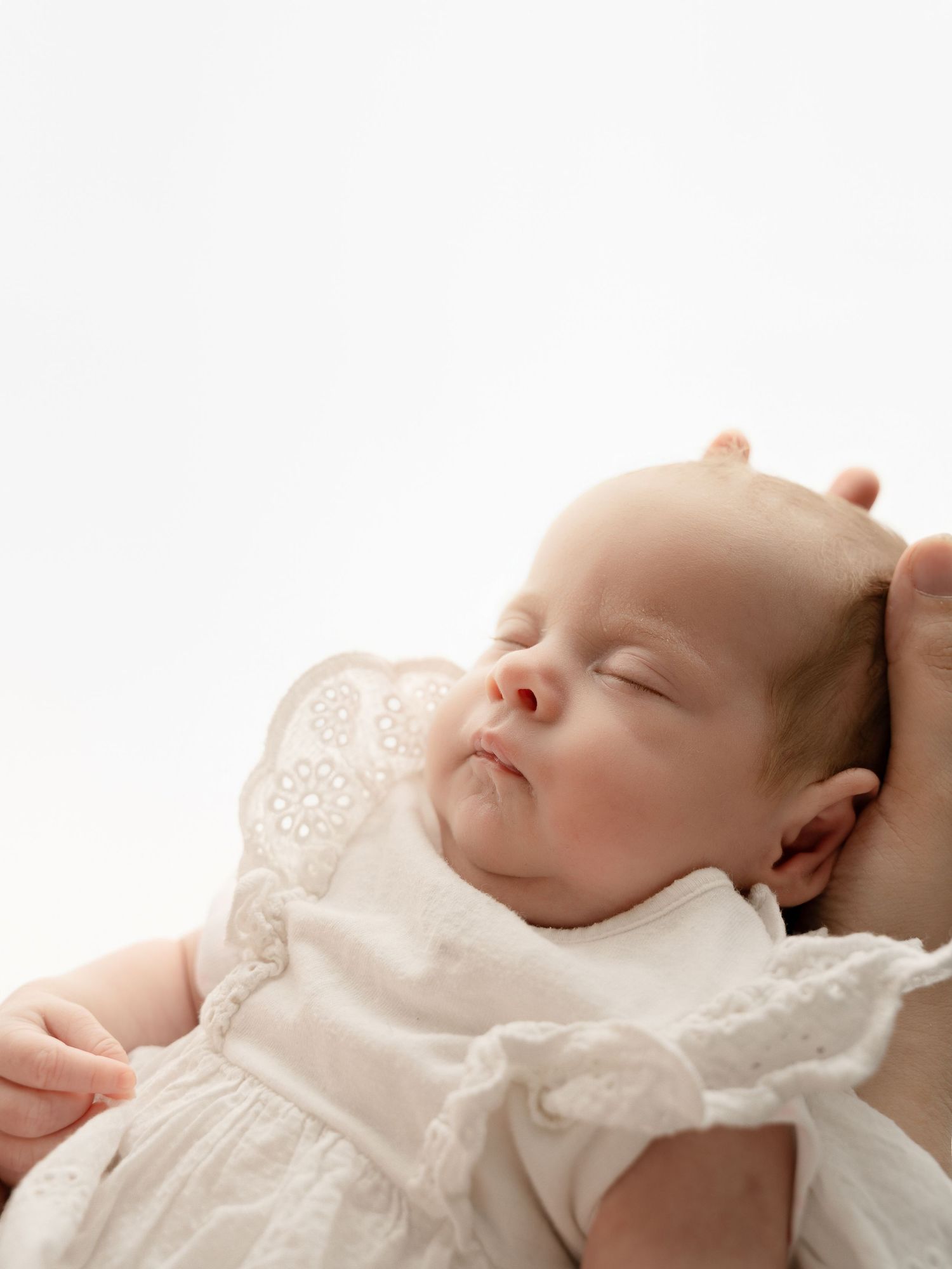 Sleeping newborn baby in white eyelet dress held in arms.