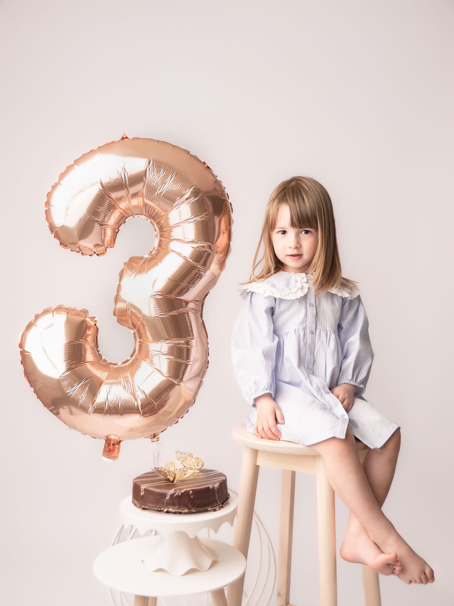 A little girl sits on a stool beside a chocolate cake and rose gold number 3 balloon for her birthday.
