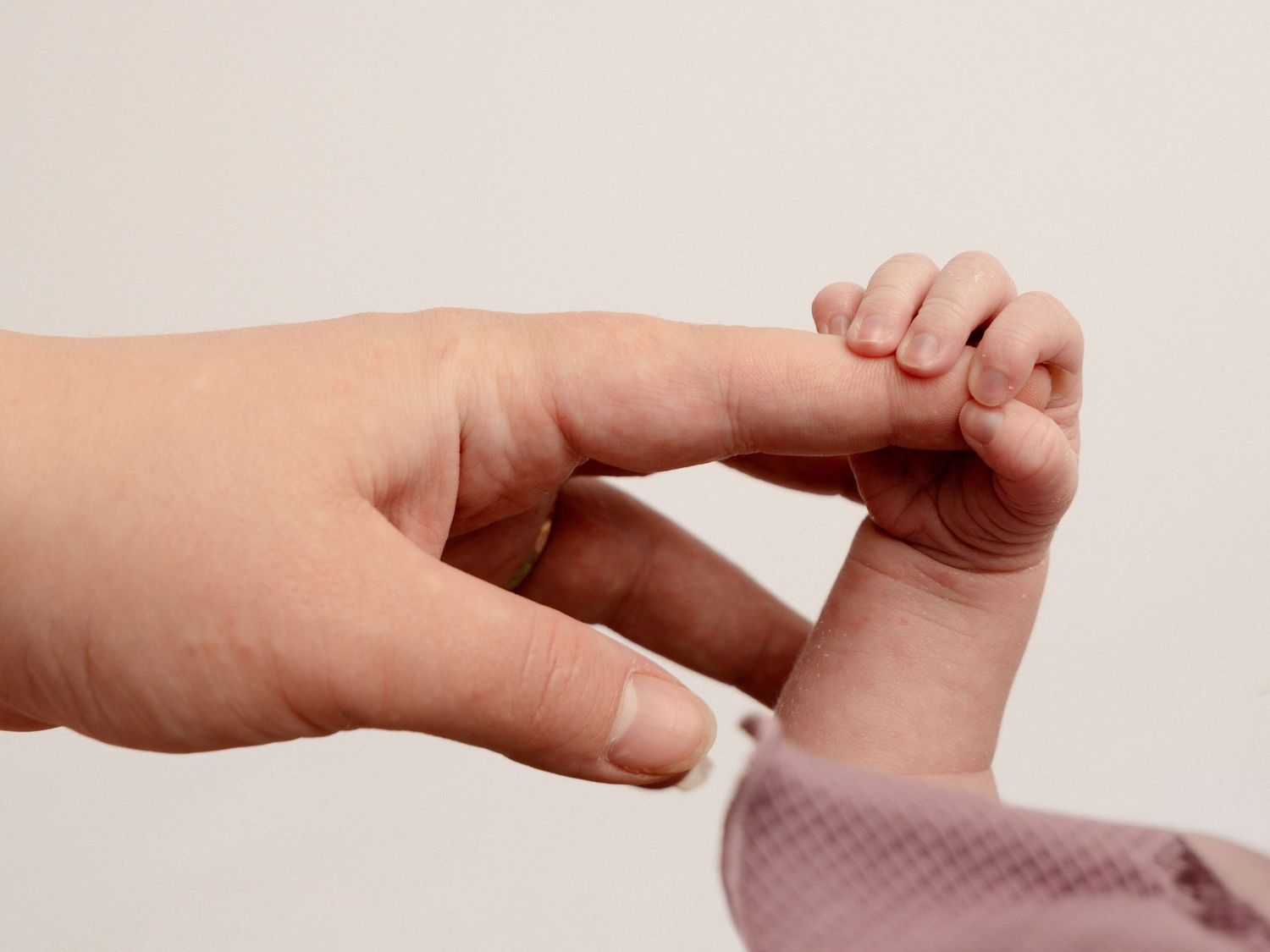 Newborn baby tiny hand grasping adult finger against light gray background.