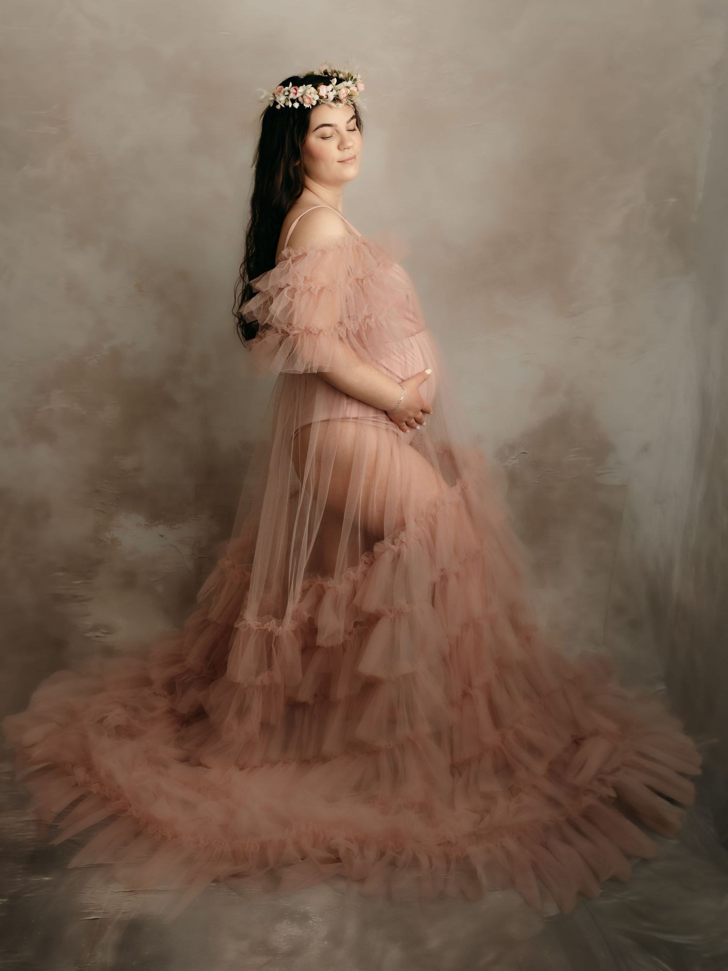 Pregnant woman in a blush pink ruffled tulle gown with a floral crown posing for a maternity portrait.