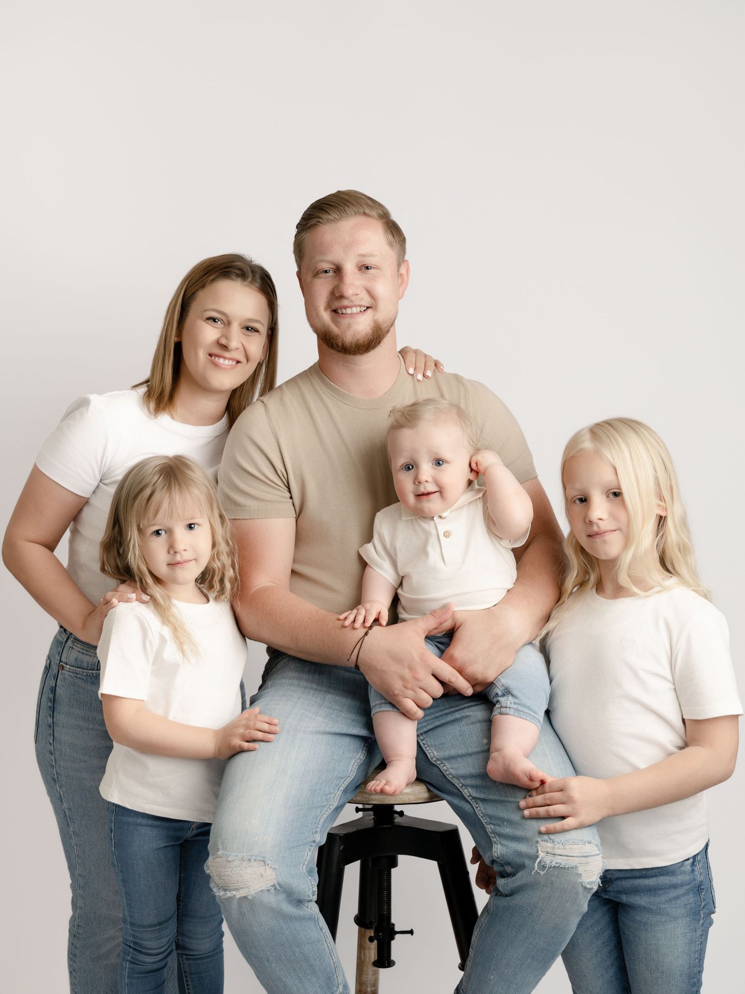 A family of five in matching white tops and jeans poses together in a bright studio portrait.