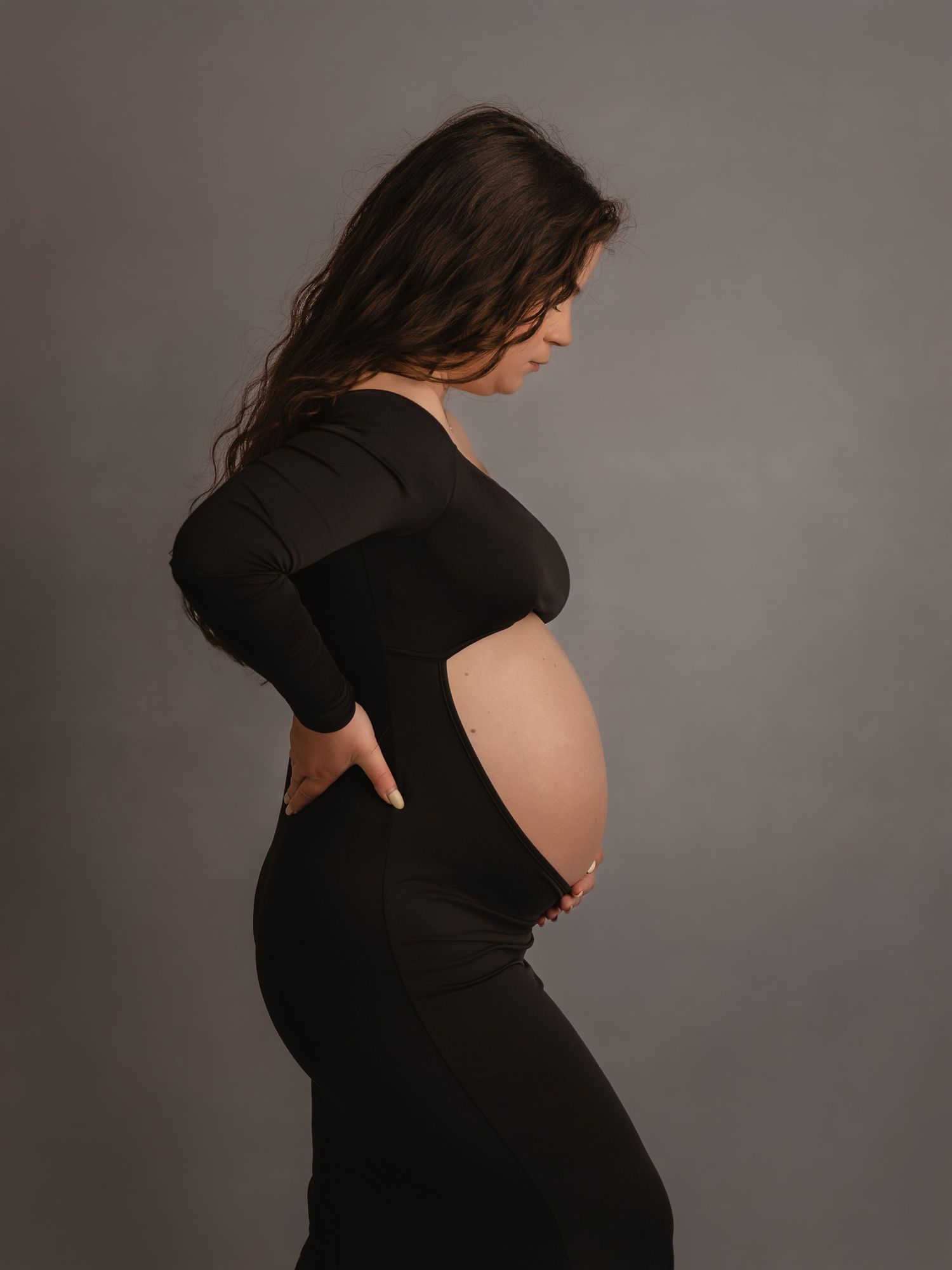 Pregnant woman in black off-shoulder maternity dress showing bare baby bump in side profile studio portrait.