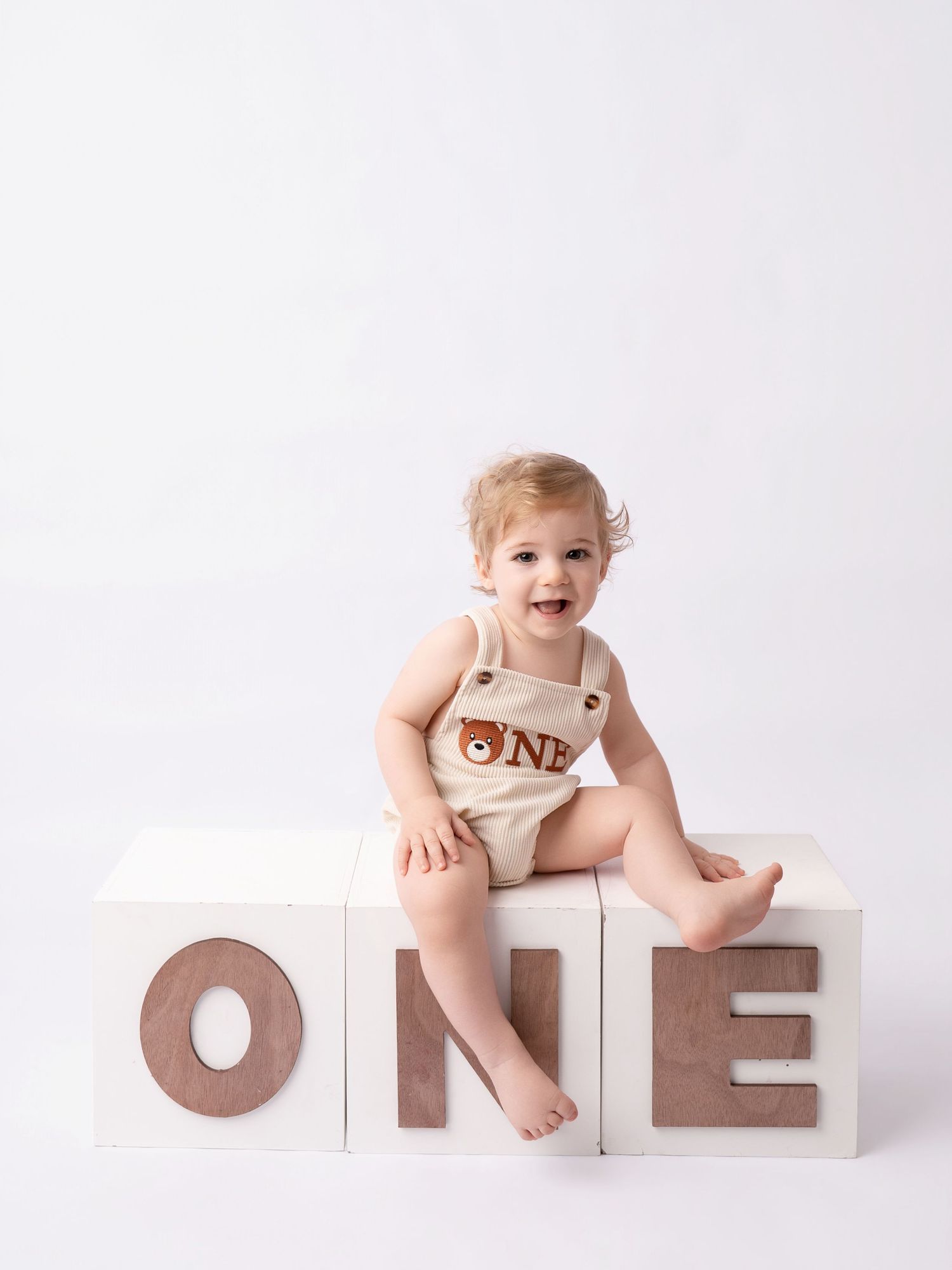 Smiling baby in bear romper sits on white blocks spelling ONE for a first birthday photo session.