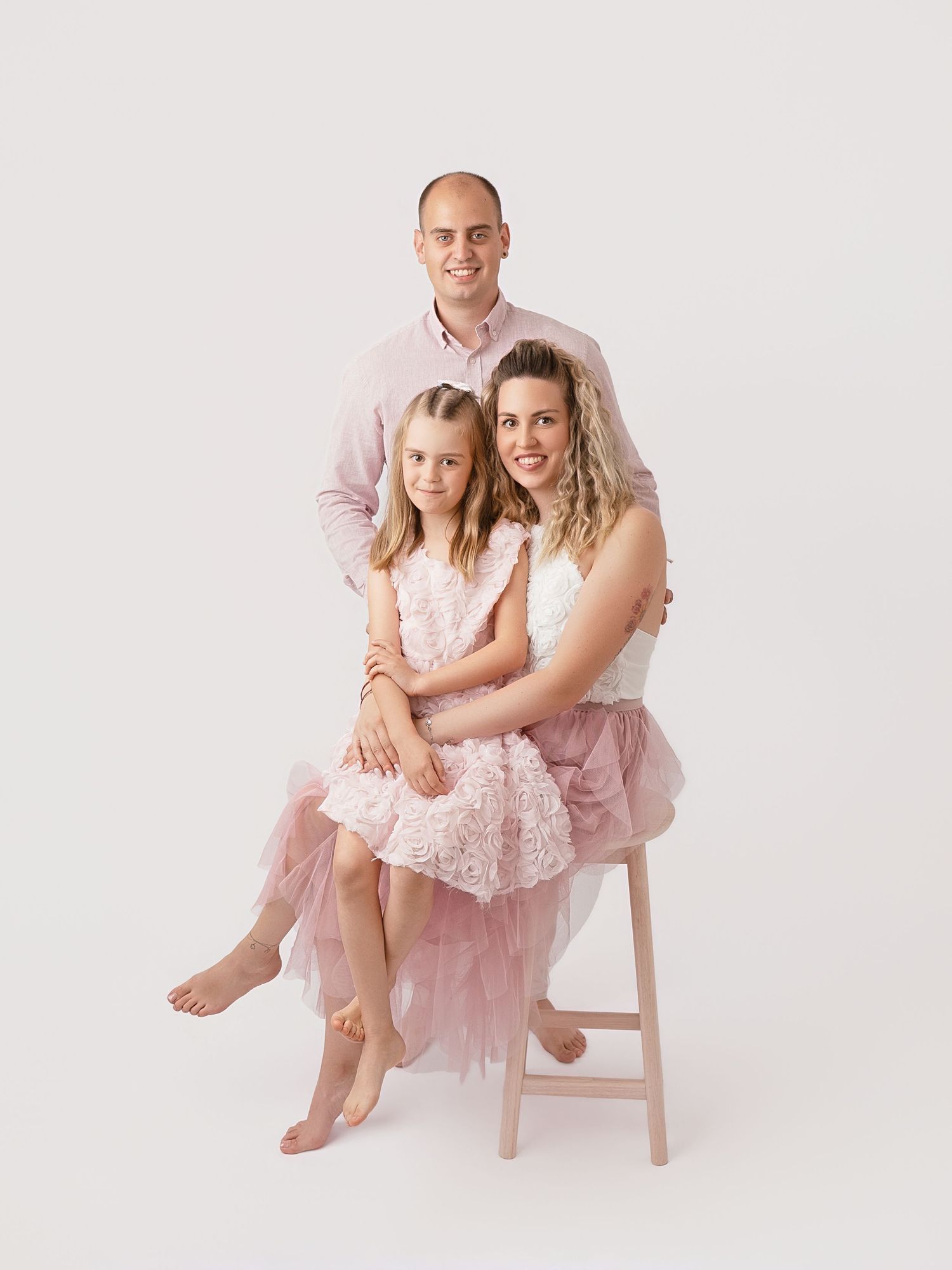 Family portrait of man, woman, and young girl in pink outfits posing together in a bright studio setting.